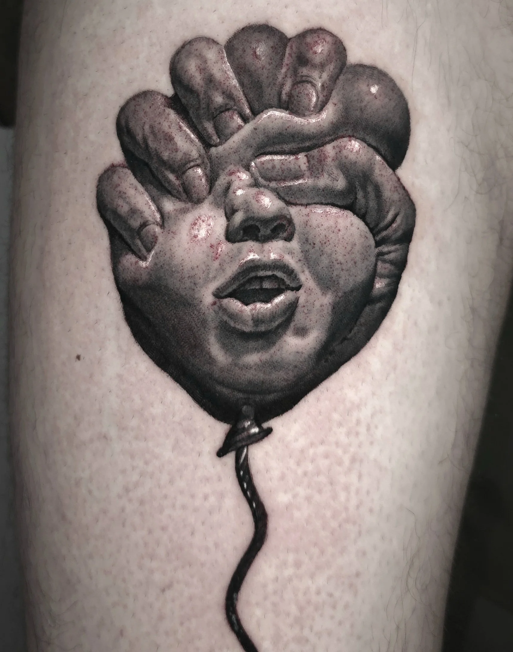 A tattoo of a balloon with a realistic face and hands, with the hands covering the eyes and the mouth open. The balloon string is attached at the bottom.