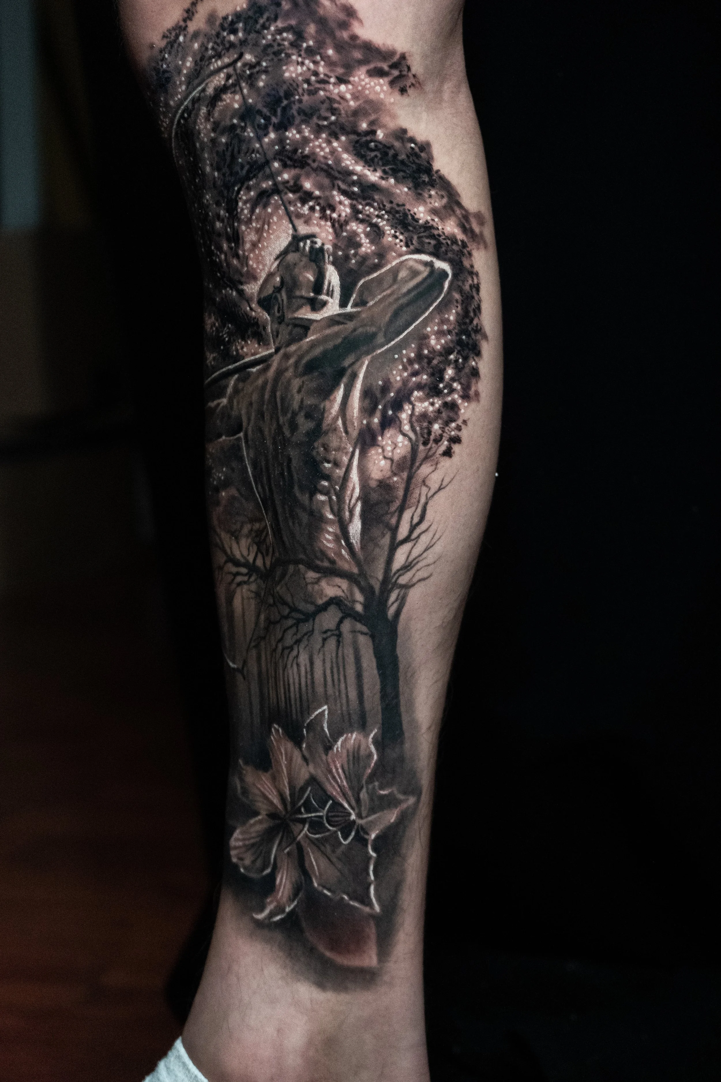 Black and gray tattoo of a hand holding a key, with trees and a butterfly in a natural scene.
