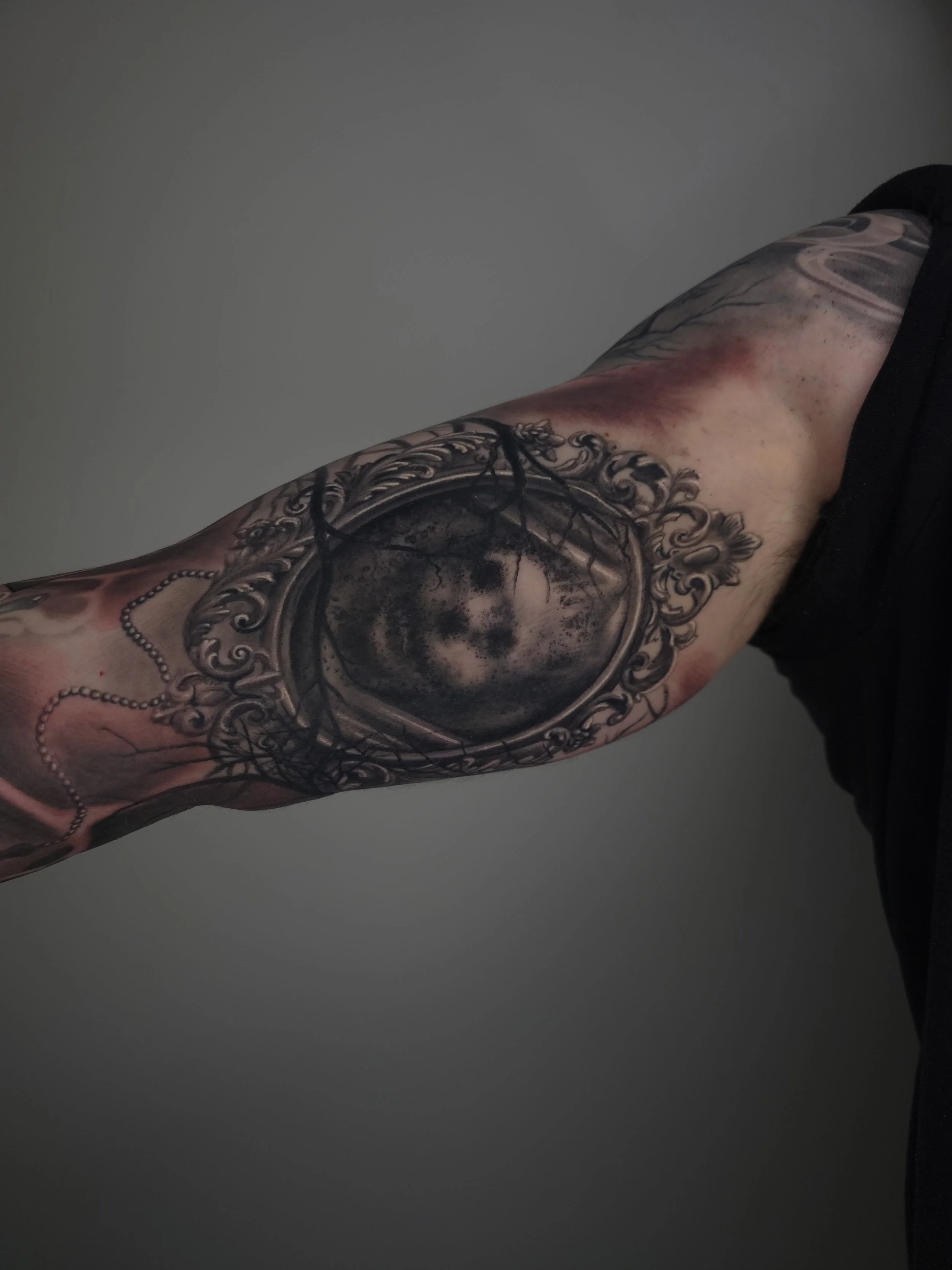 Close-up of a person's arm with an intricate, black and grey tattoo of a vintage pocket mirror with a woman's face reflection, surrounded by ornate floral and decorative elements.