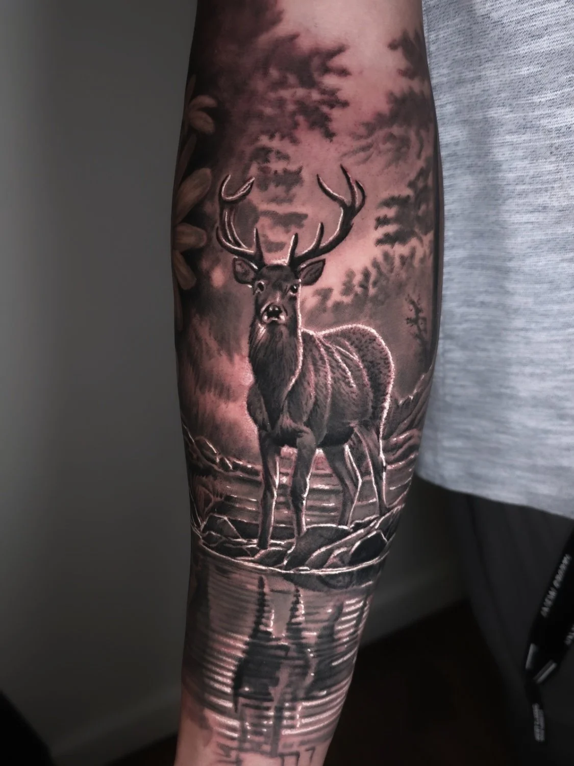 Black and gray tattoo of a buck standing at the edge of a river, with a cloudy sky and trees in the background.