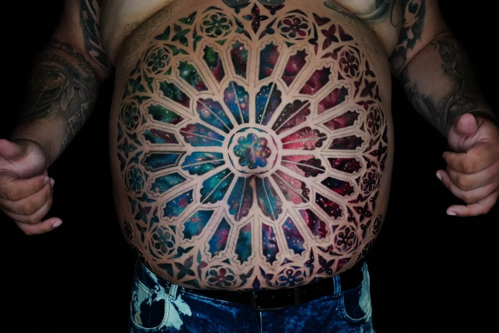 A person's bare chest with a large, elaborate tattoo resembling a colorful stained glass window featuring a galaxy pattern of stars, nebulae, and cosmic colors, surrounded by intricate black linework. The person is holding their arms out.