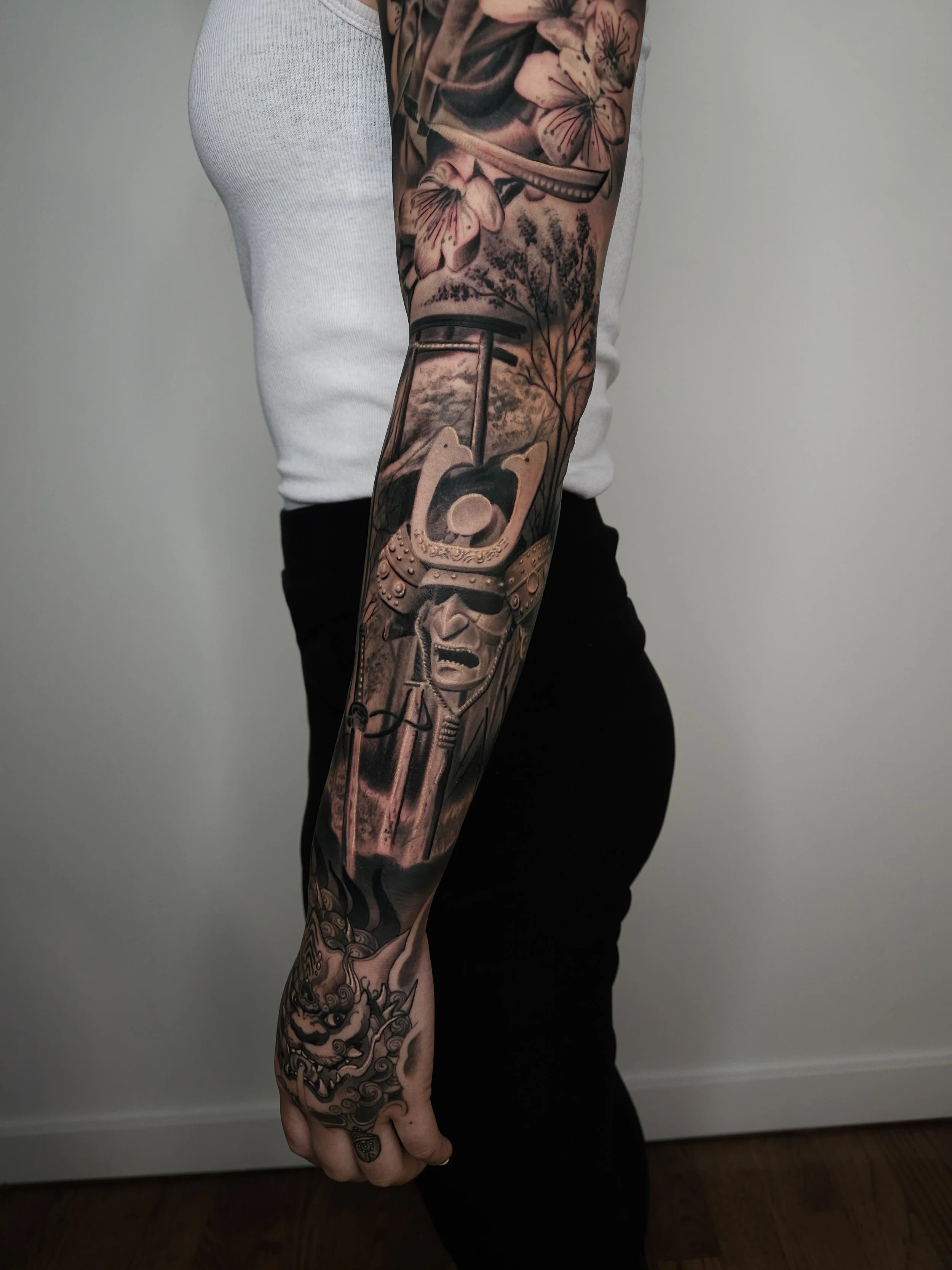 Tattooed person's arm displaying a sleeve tattoo with various designs including flowers, a sword, a mask, and a demon face.