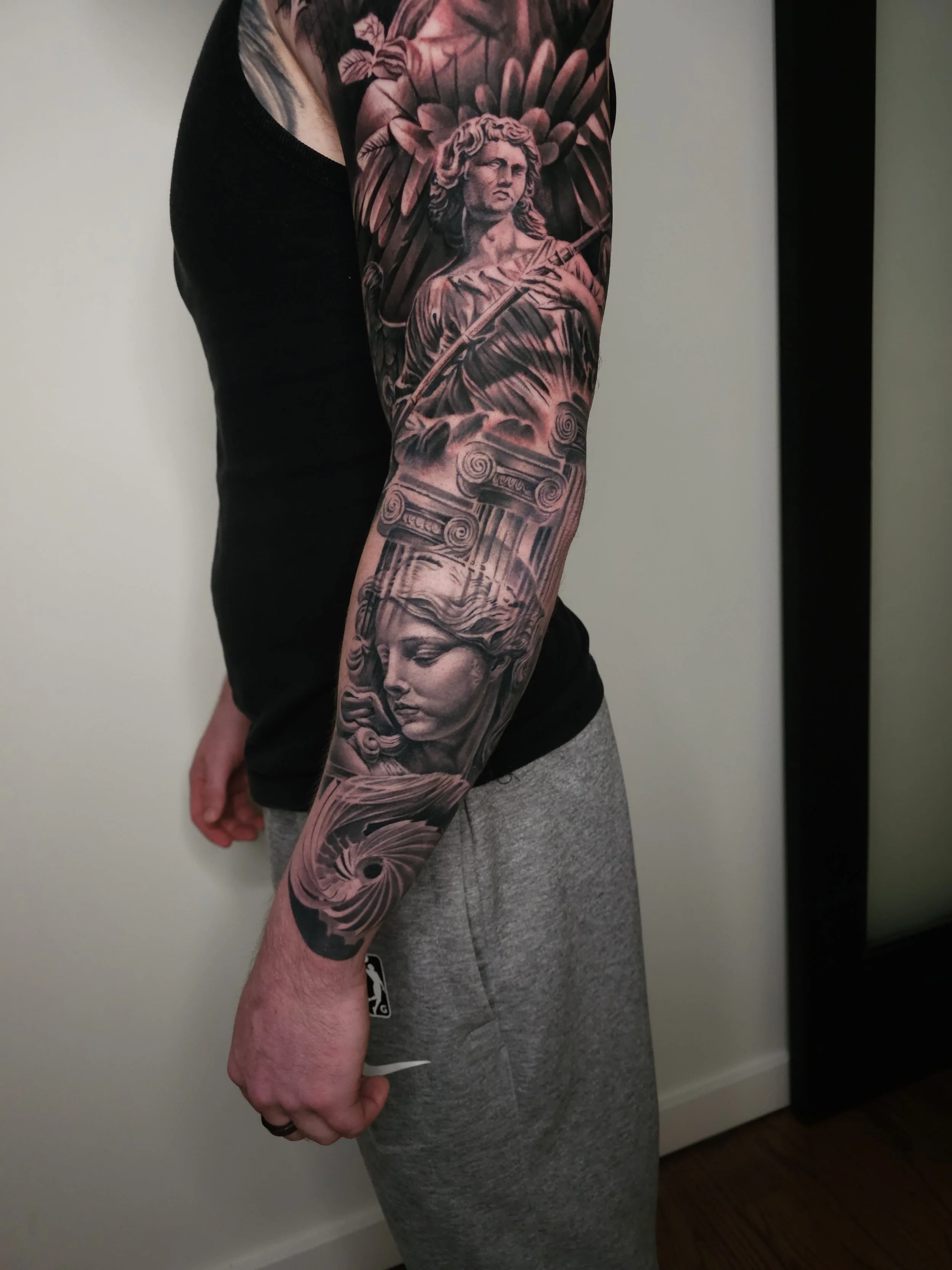 A person's arm with a detailed black and grey tattoo sleeve featuring classical sculptures and elements from Greek mythology, including a statue of a woman with flowing hair and a figure with wings surrounded by floral patterns.