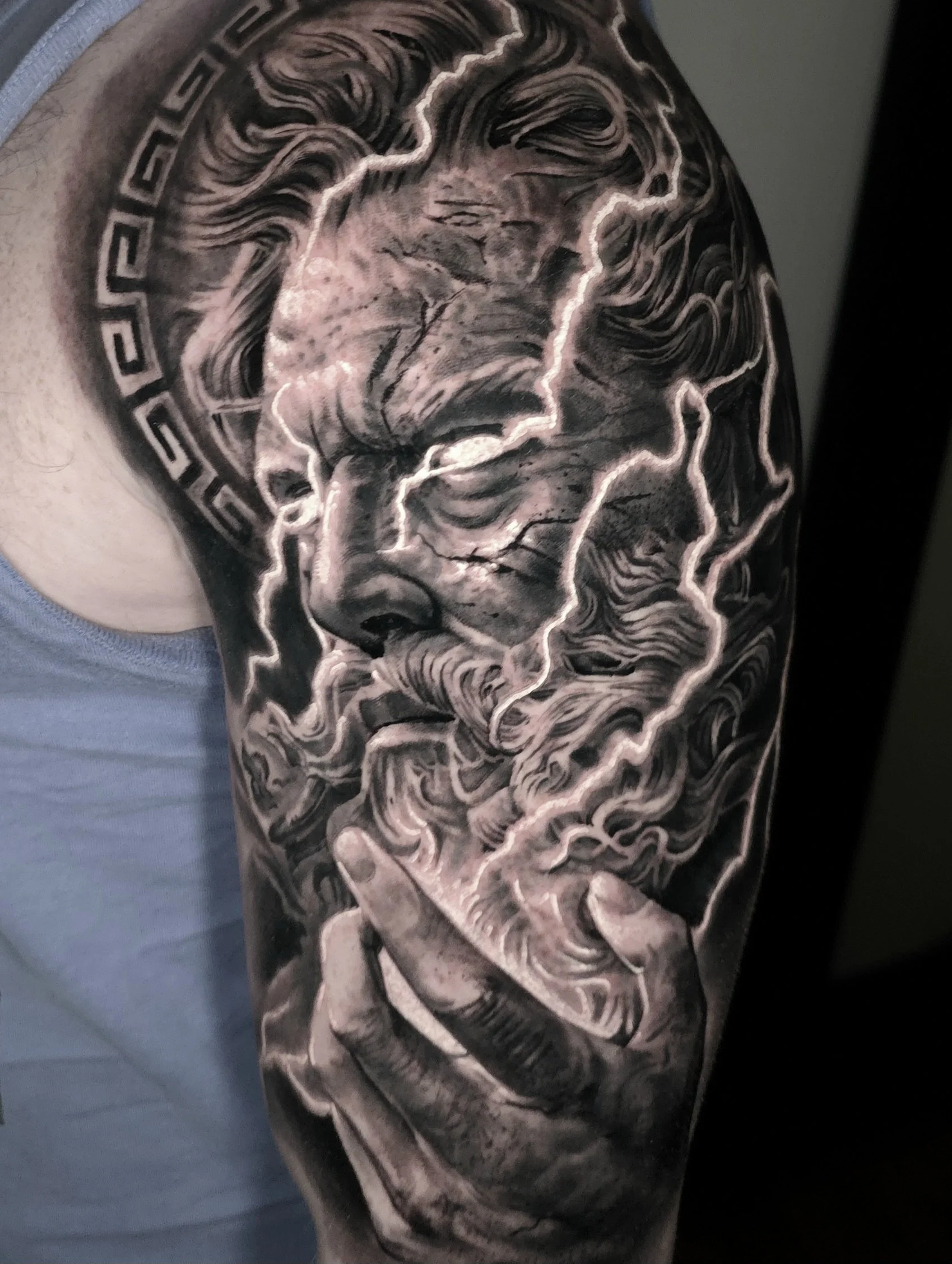 Black and gray tattoo of a classical statue's face with flowing hair, surrounded by lightning bolts and a Greek key border.