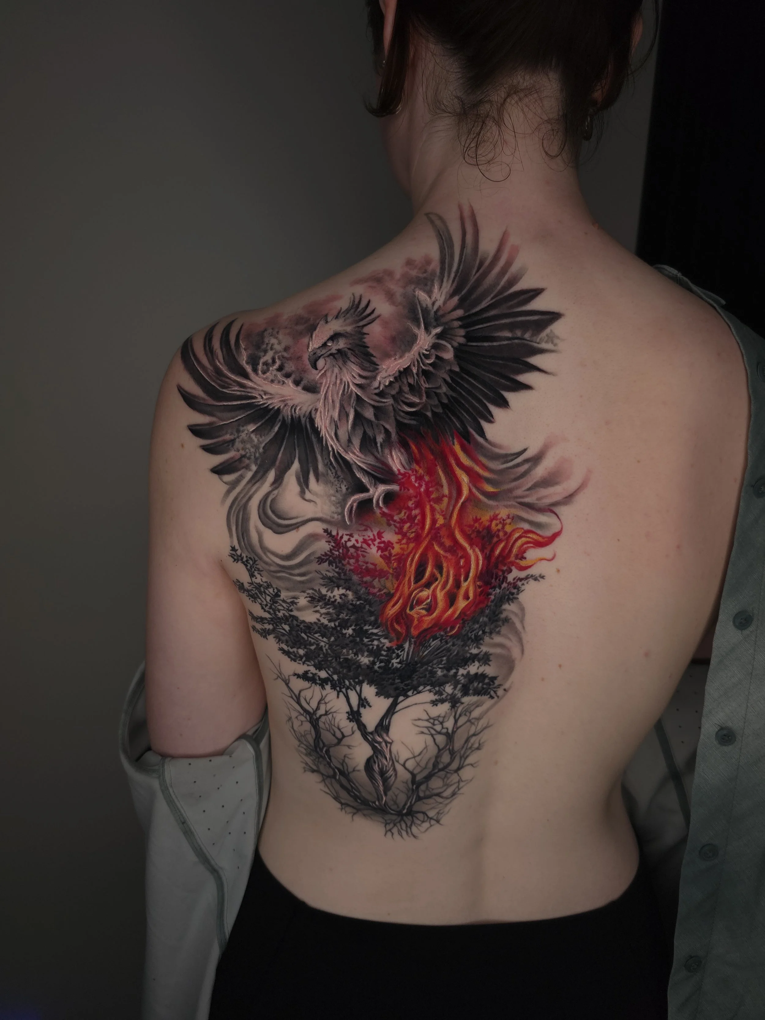 A tattoo on a person's back featuring a phoenix rising above a fiery tree with leafless branches.