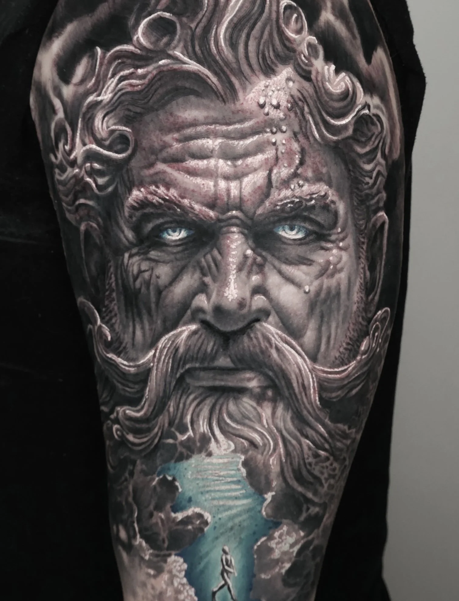 Tattoo of a bearded man with long flowing hair, piercing blue eyes, and a serious expression, with a scene of a person walking towards a lighted tunnel in the lower part.