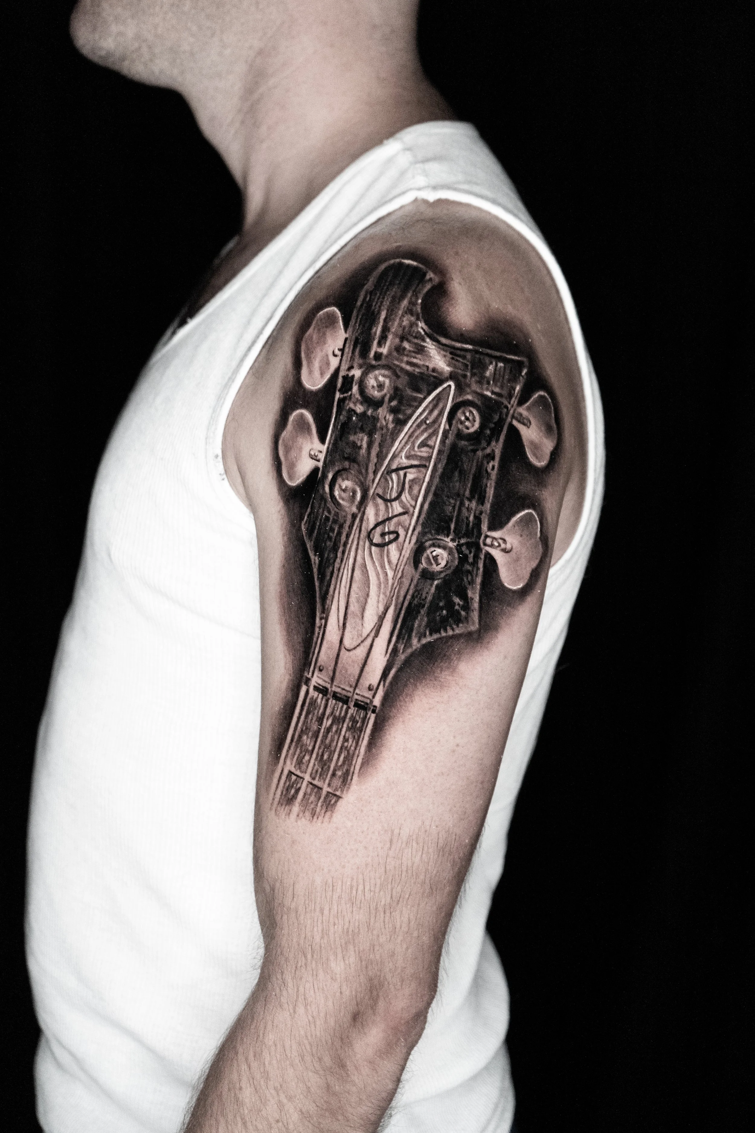 A person's upper arm with a realistic black and gray tattoo of a guitar headstock on the shoulder.