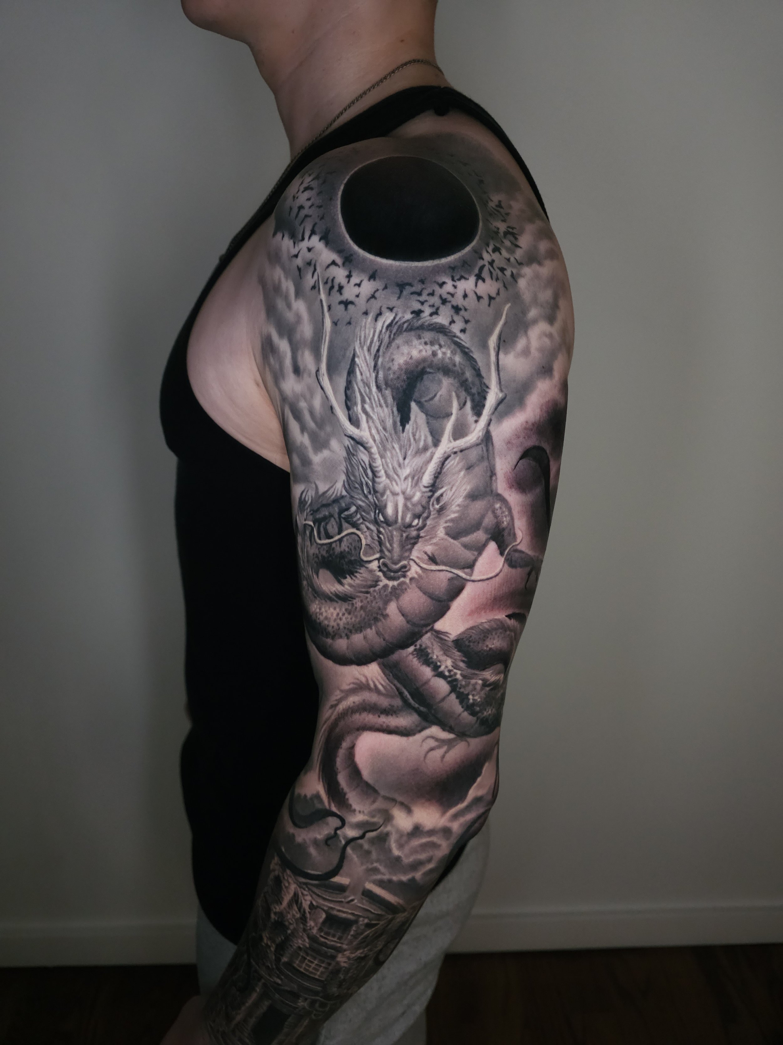 Black and gray tattoo of a dragon with antlers on a person's upper arm and shoulder, featuring a dark moon and clouds in the background.
