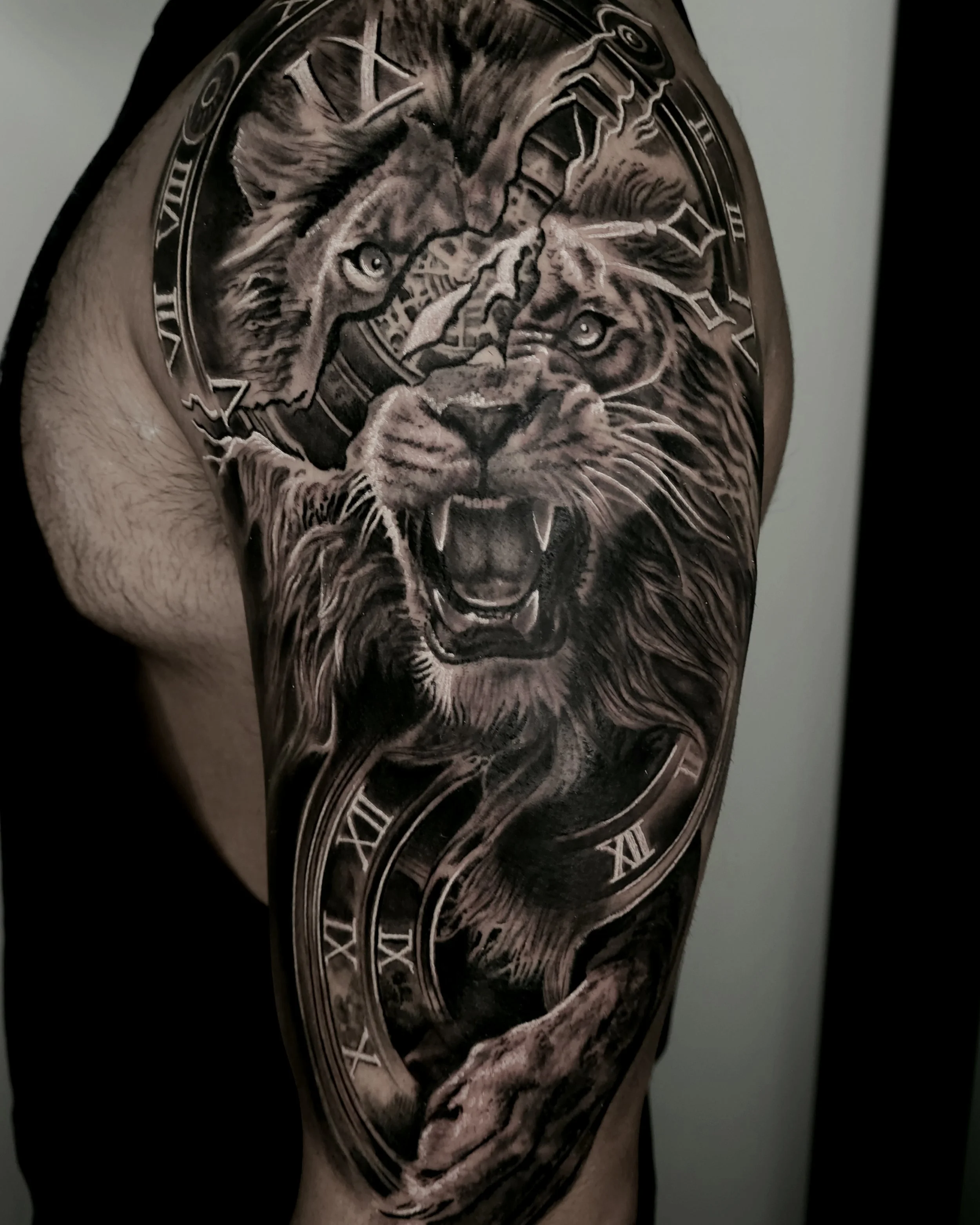 Black and gray tattoo on upper arm featuring a roaring lion's face in the foreground, with a cub's face behind it, a clock with Roman numerals, and swirling decorative elements.