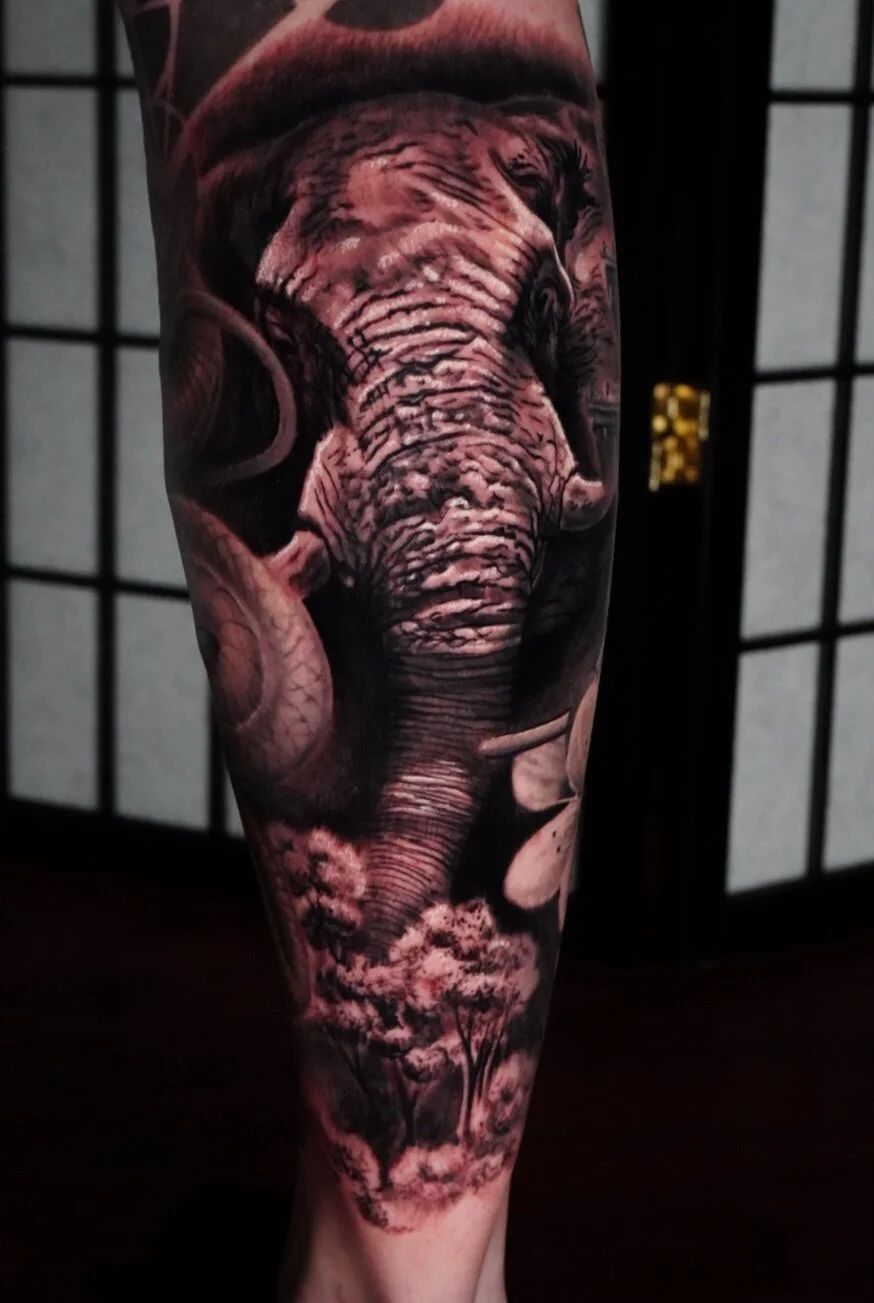 Tattoo of an elephant's face and trunk on a person's leg, with detailed shading of trees and background elements.