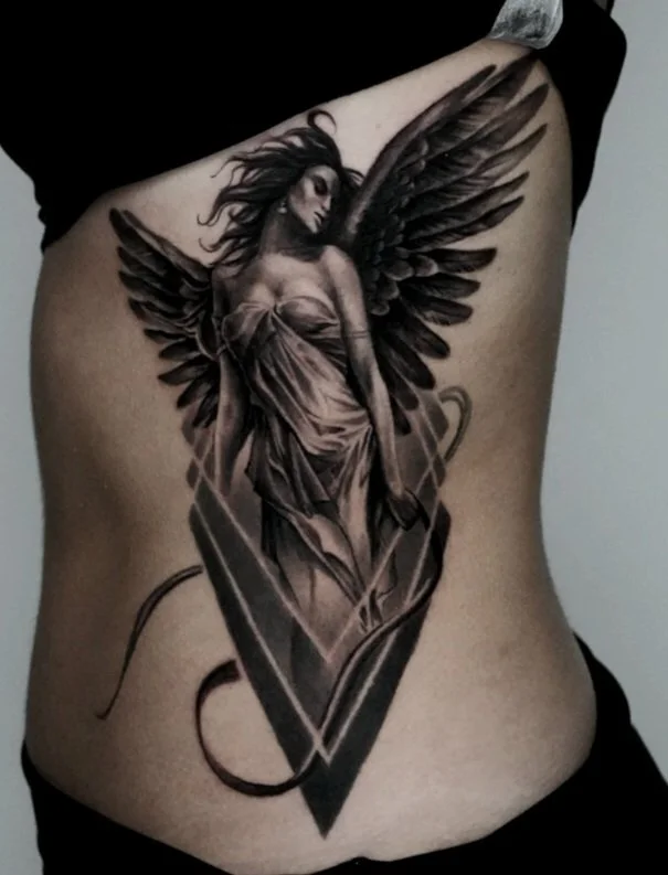 A black and gray tattoo of a woman with wings, standing with her head tilted to the side and hair flowing, on a person's side torso.