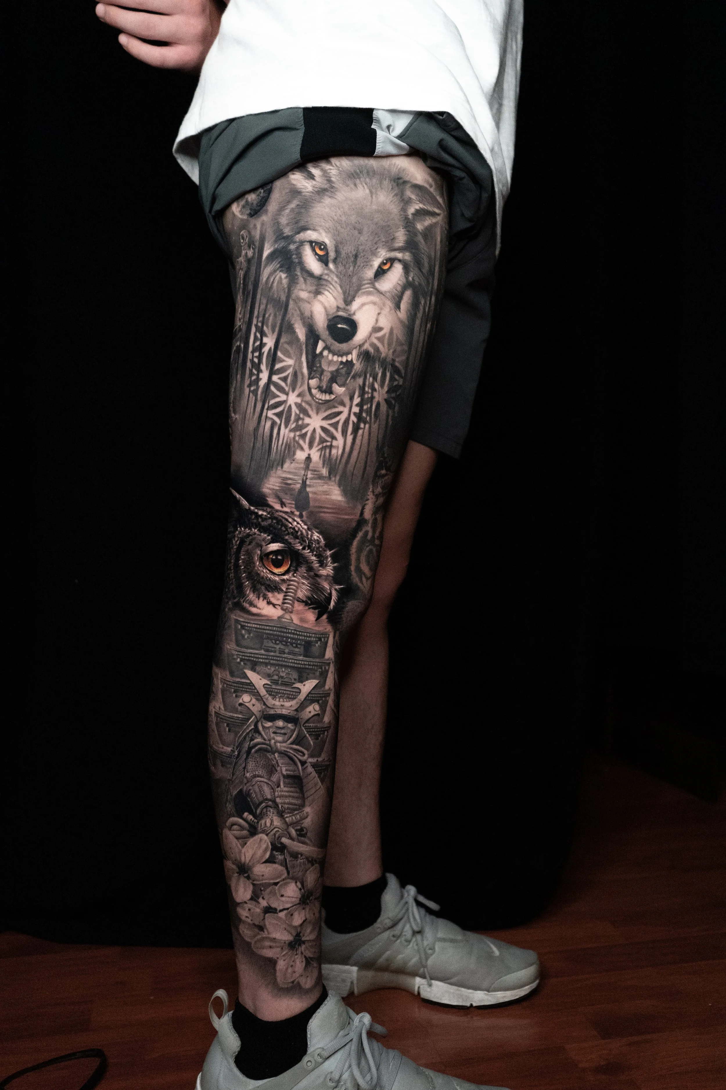 A person with a tattoo sleeve on their leg featuring a snarling wolf, an owl, a samurai, and flowers.