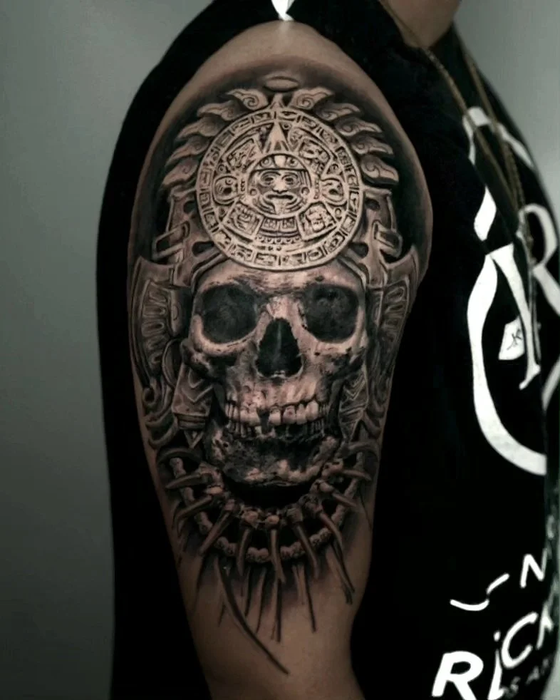 Black and gray tattoo of a human skull with intricate Aztec designs and a sun motif on top.
