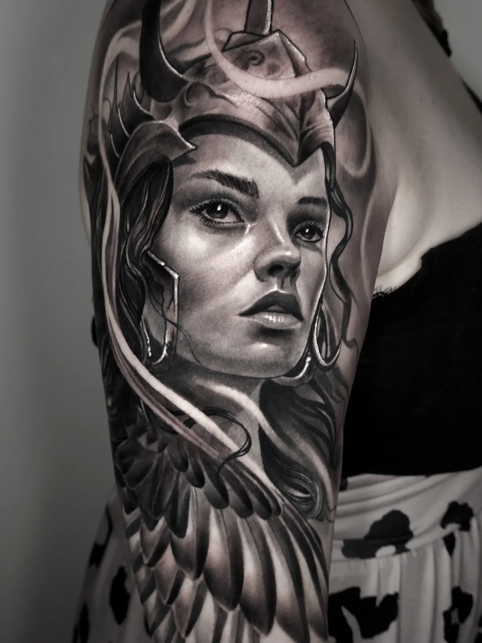 Black and gray realistic tattoo of a woman's face with detailed hair, earrings, and a headdress, surrounded by feather-like designs on person's upper arm.