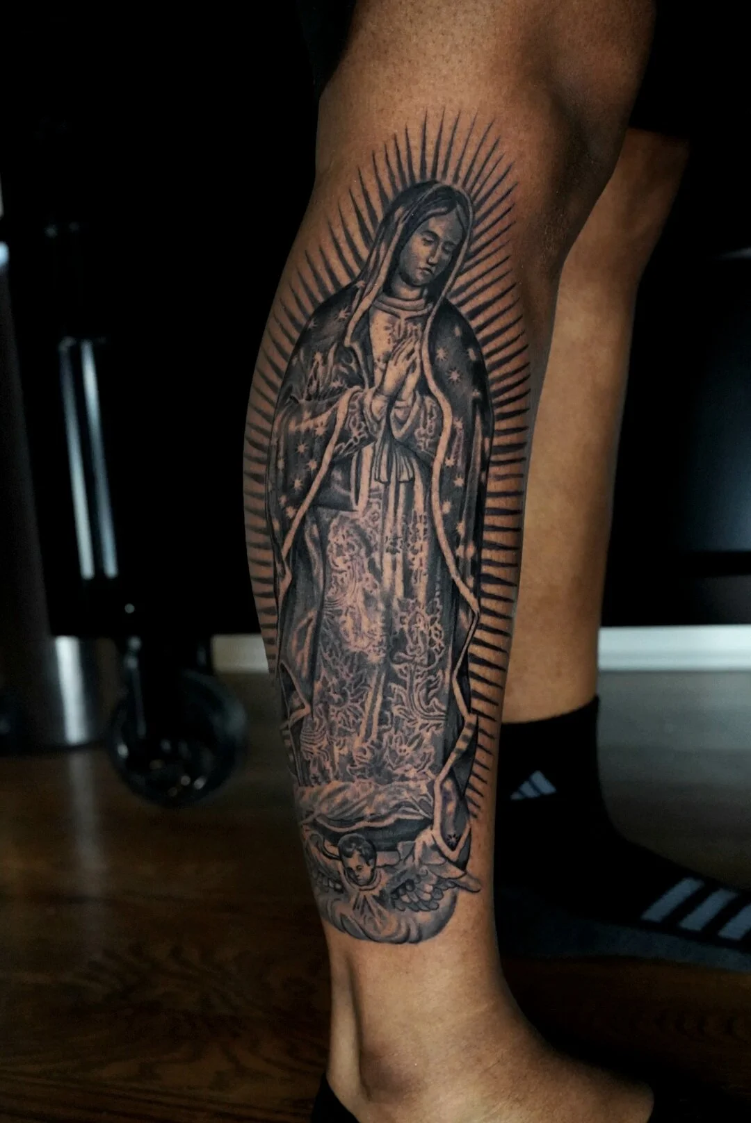 Black and gray tattoo of the Virgin Mary with a halo, hands in prayer, and a detailed robe, located on the person's forearm.