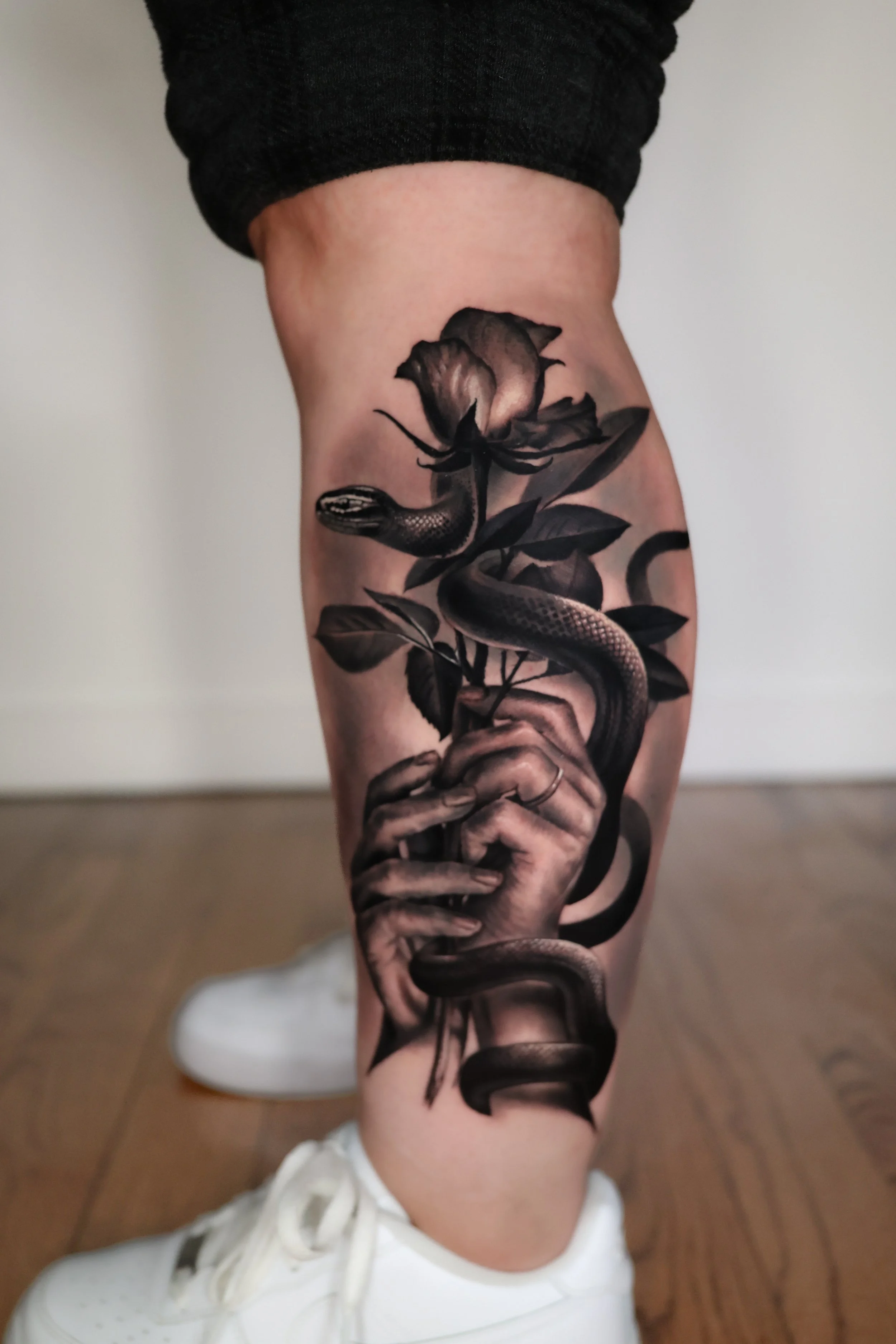 A black and gray tattoo on a person's leg featuring a snake intertwined with a hand holding a rose and leaves.