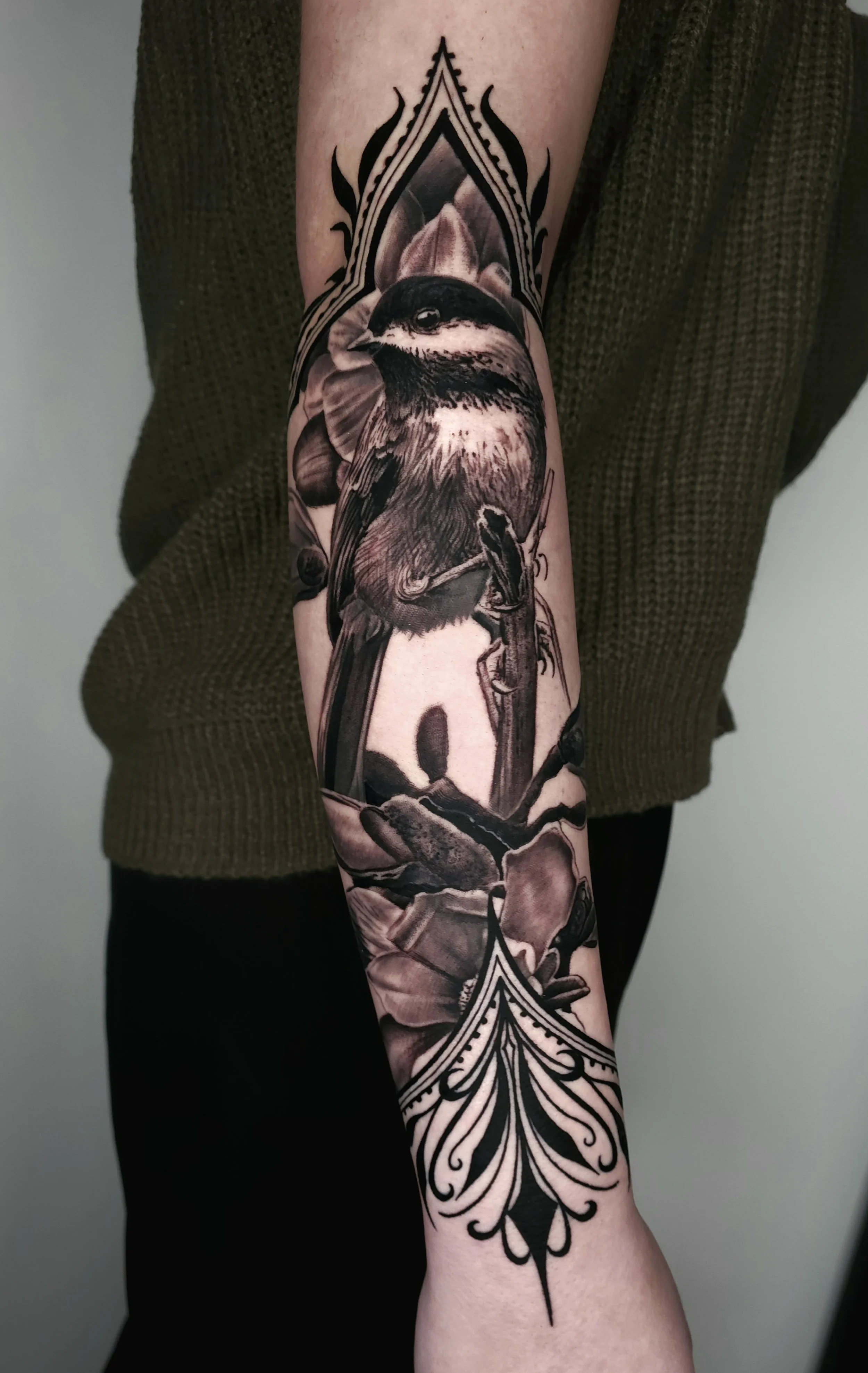 Black and gray realism tattoo of a bird perched on a branch, surrounded by leaves and floral elements, with decorative ornamental patterns at the top and bottom.