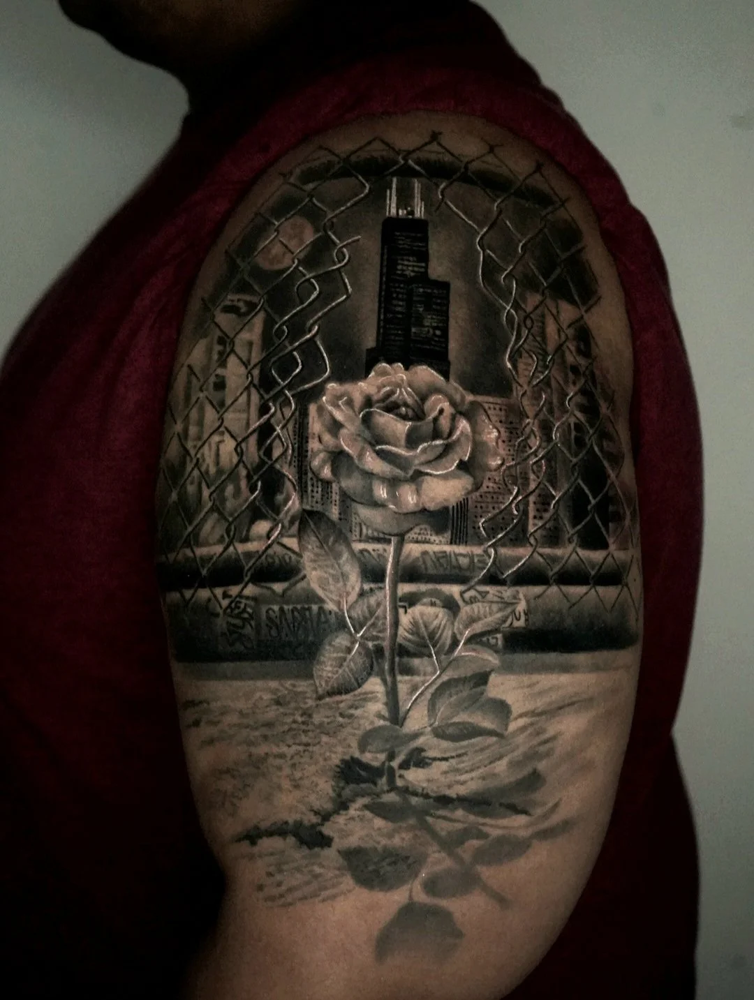 Black and gray tattoo of a cityscape with a rose growing in front, framed by chain-link fence, on person's upper arm.