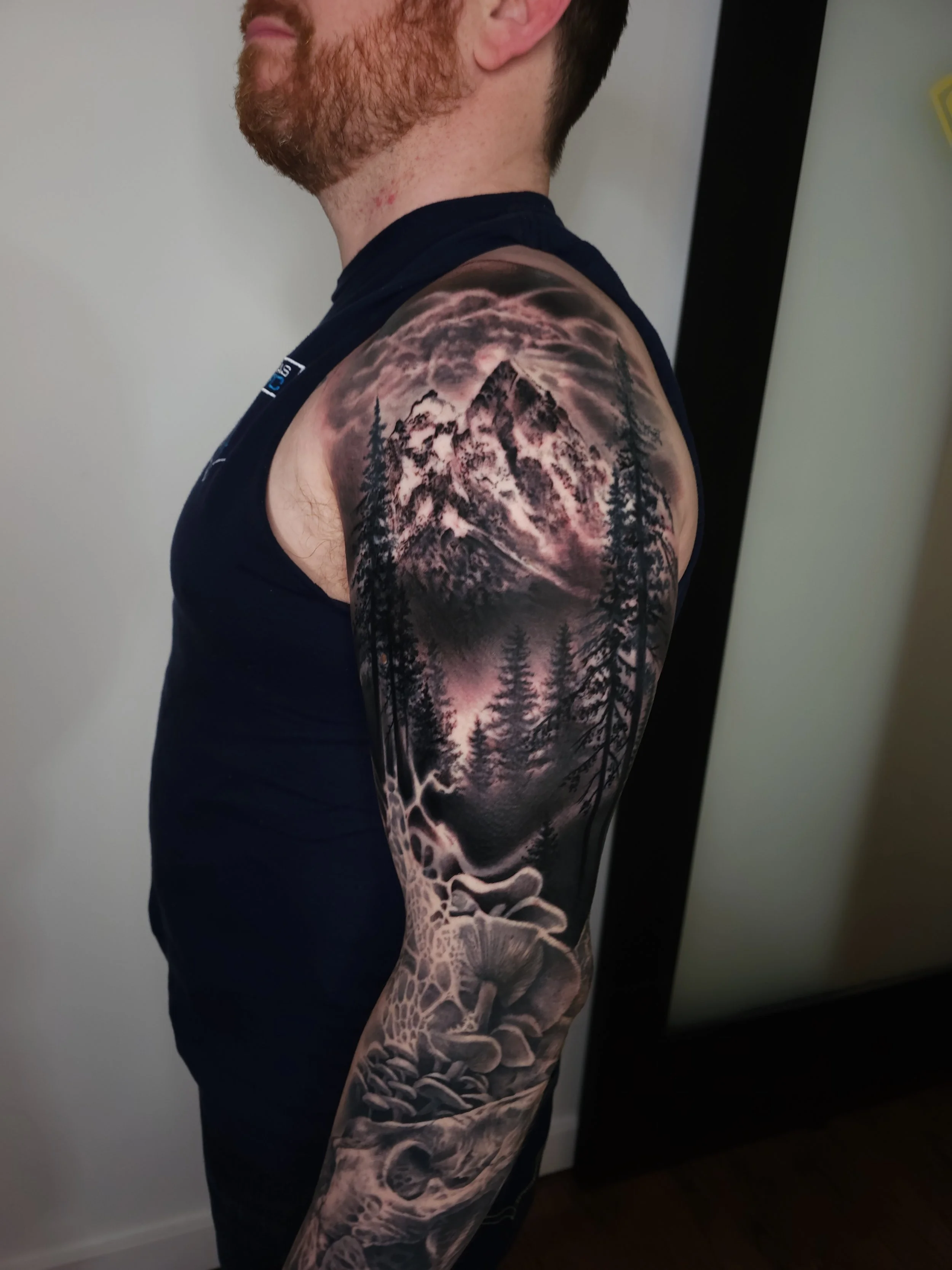 Tattoo of a mountain scene with pine trees, clouds, and mushrooms on a man's arm and shoulder.