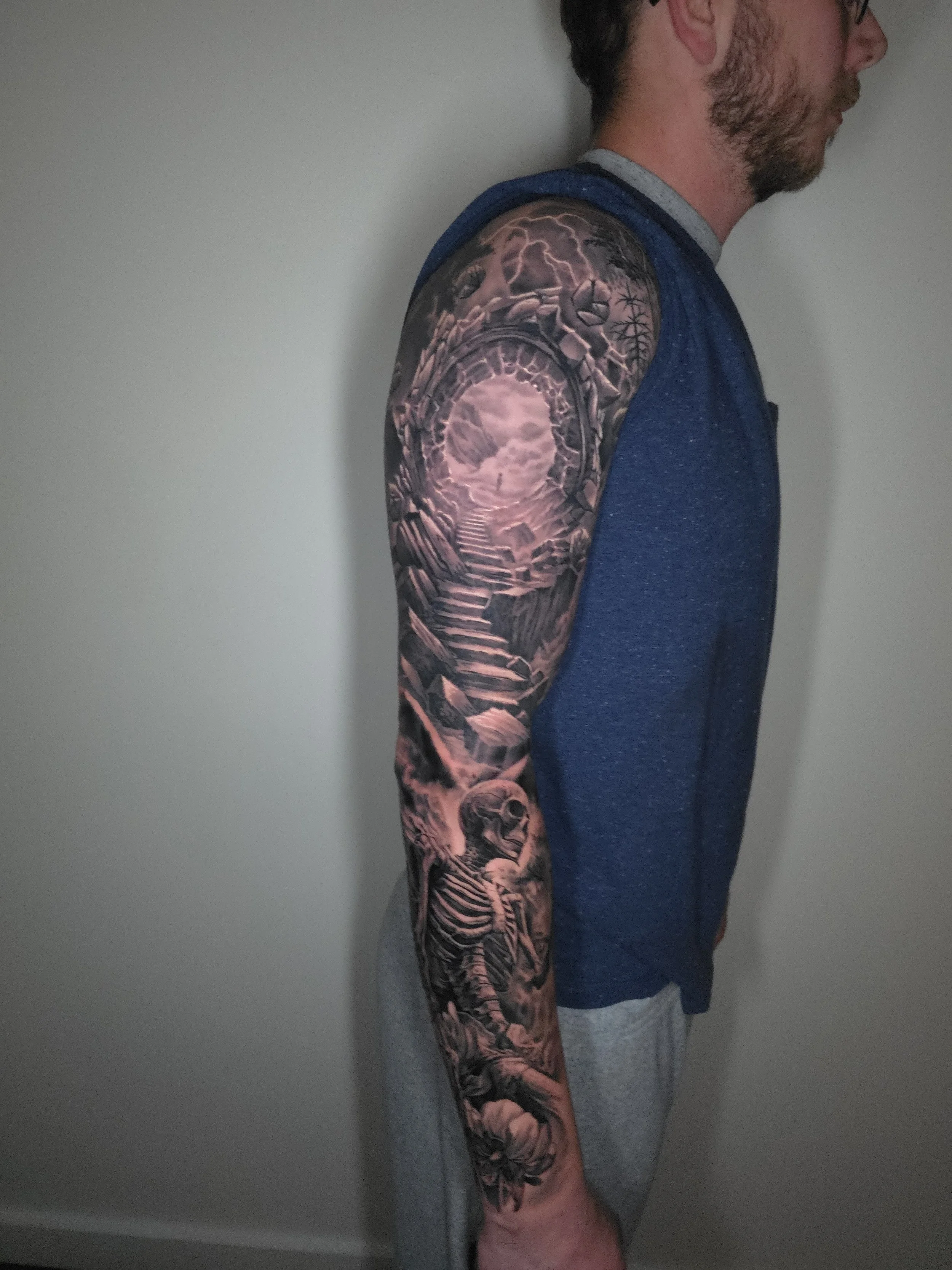 A man with a detailed black and grey sleeve tattoo on his arm, featuring imagery like a staircase, skeleton, and clouds, standing against a plain wall.