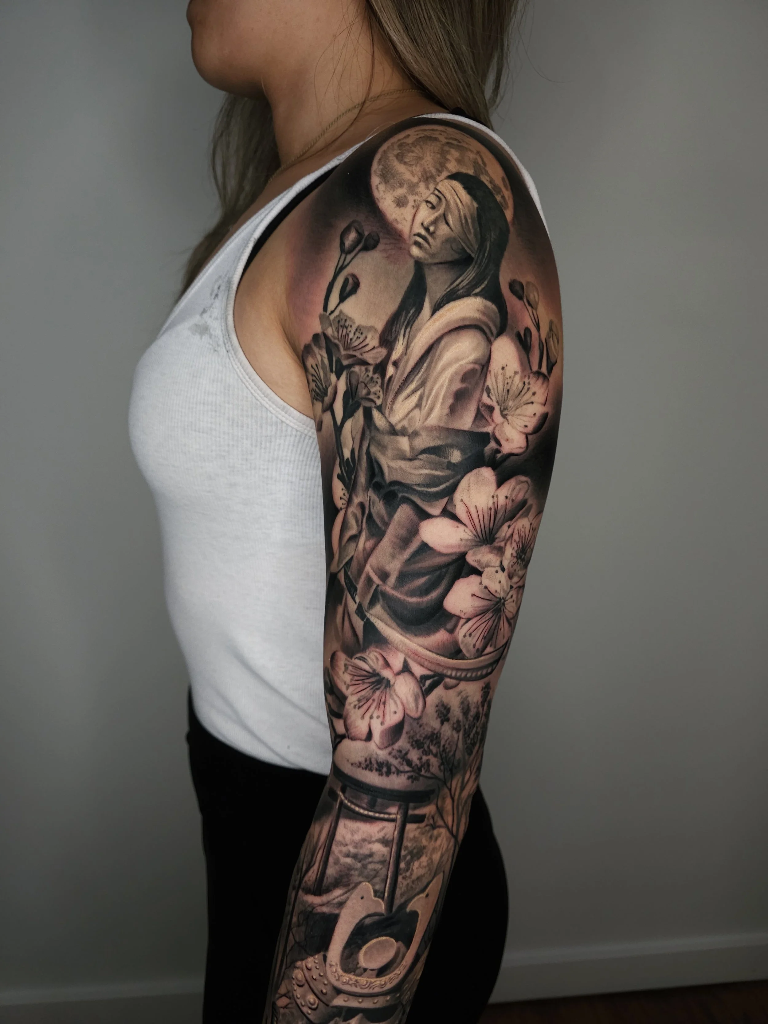 Tattoo sleeve on a woman's arm featuring a woman with a moon in the background, surrounded by flowers and a horseshoe.