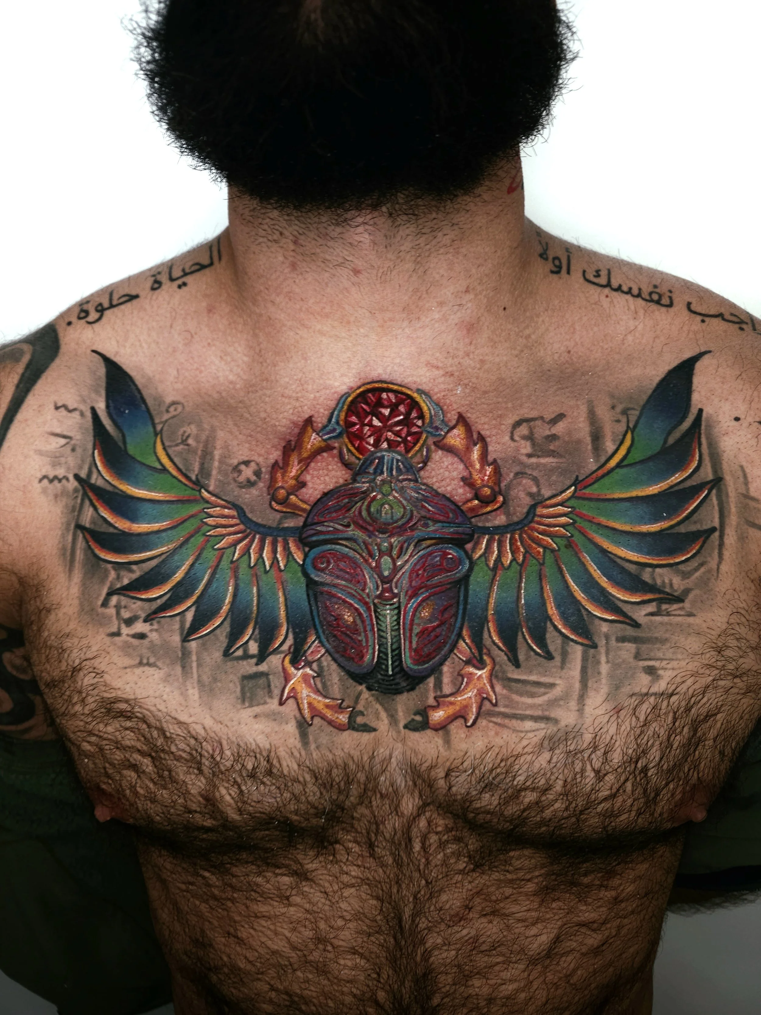 colorful tattoo of a scarab beetle with wings on man's chest, with Arabic script tattoos on shoulders.