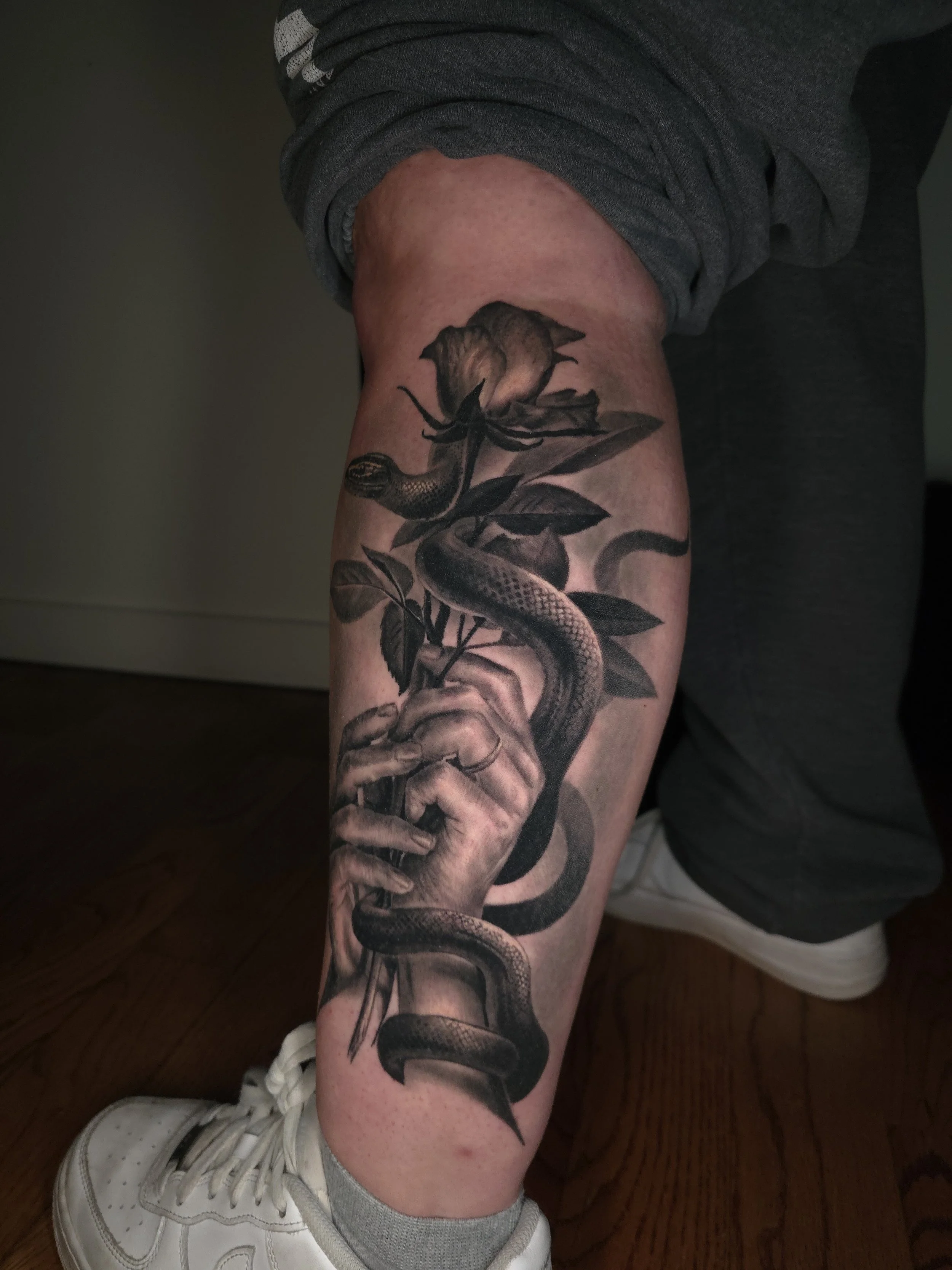 A black and gray tattoo of a hand holding a snake that wraps around a flower, located on a person's calf.