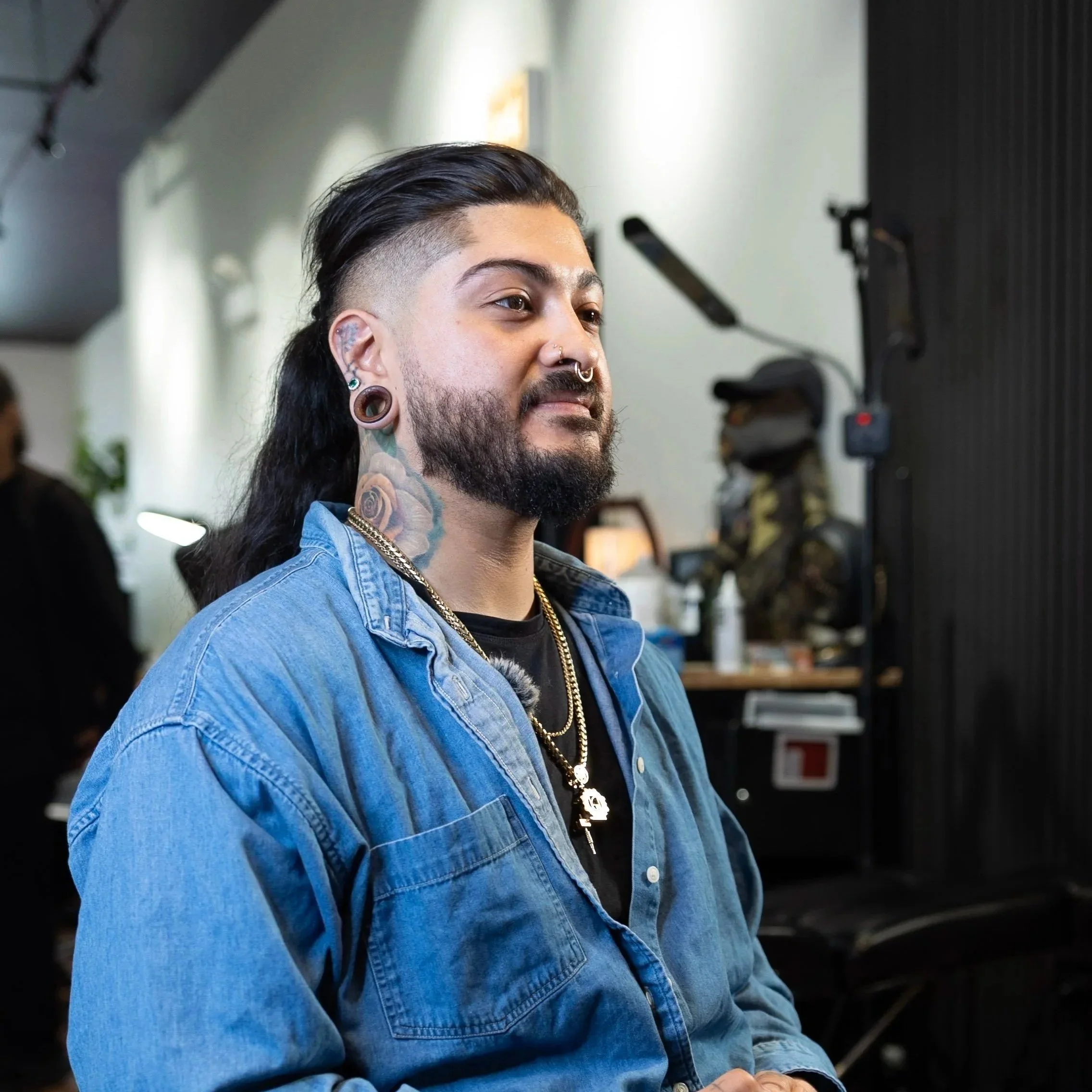 A man with long dark hair, tattoos, and pierced ears is wearing a black shirt under a blue denim jacket, looking to the side in an indoor setting with a tattoo on his neck.