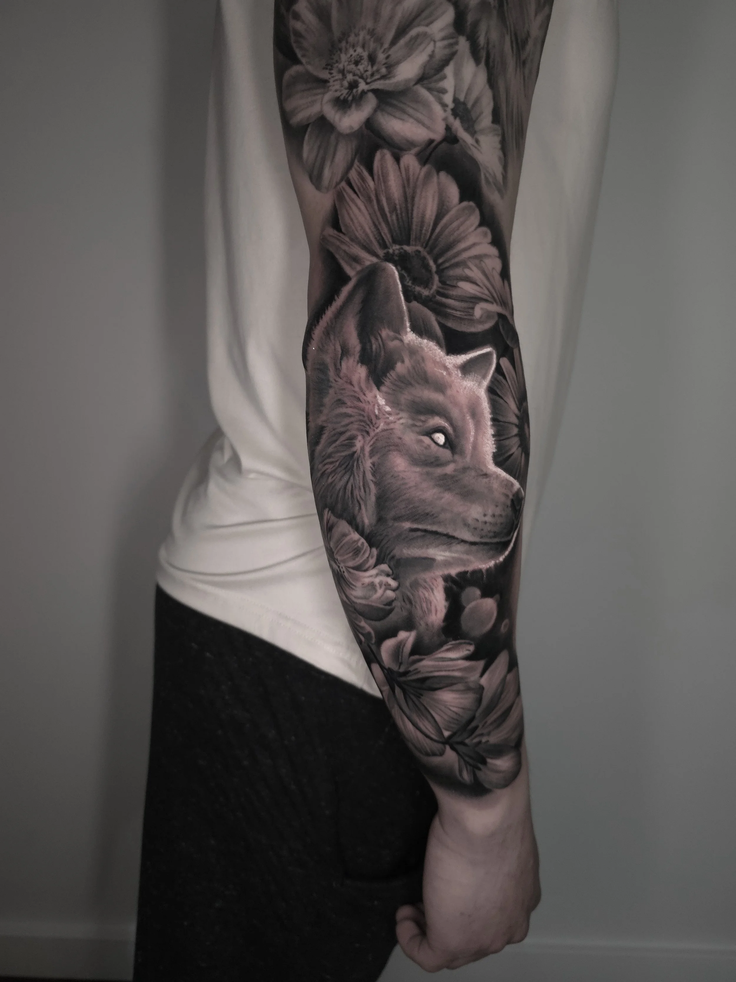 Black and gray realistic tattoo of a wolf's face surrounded by floral elements on a person's arm.