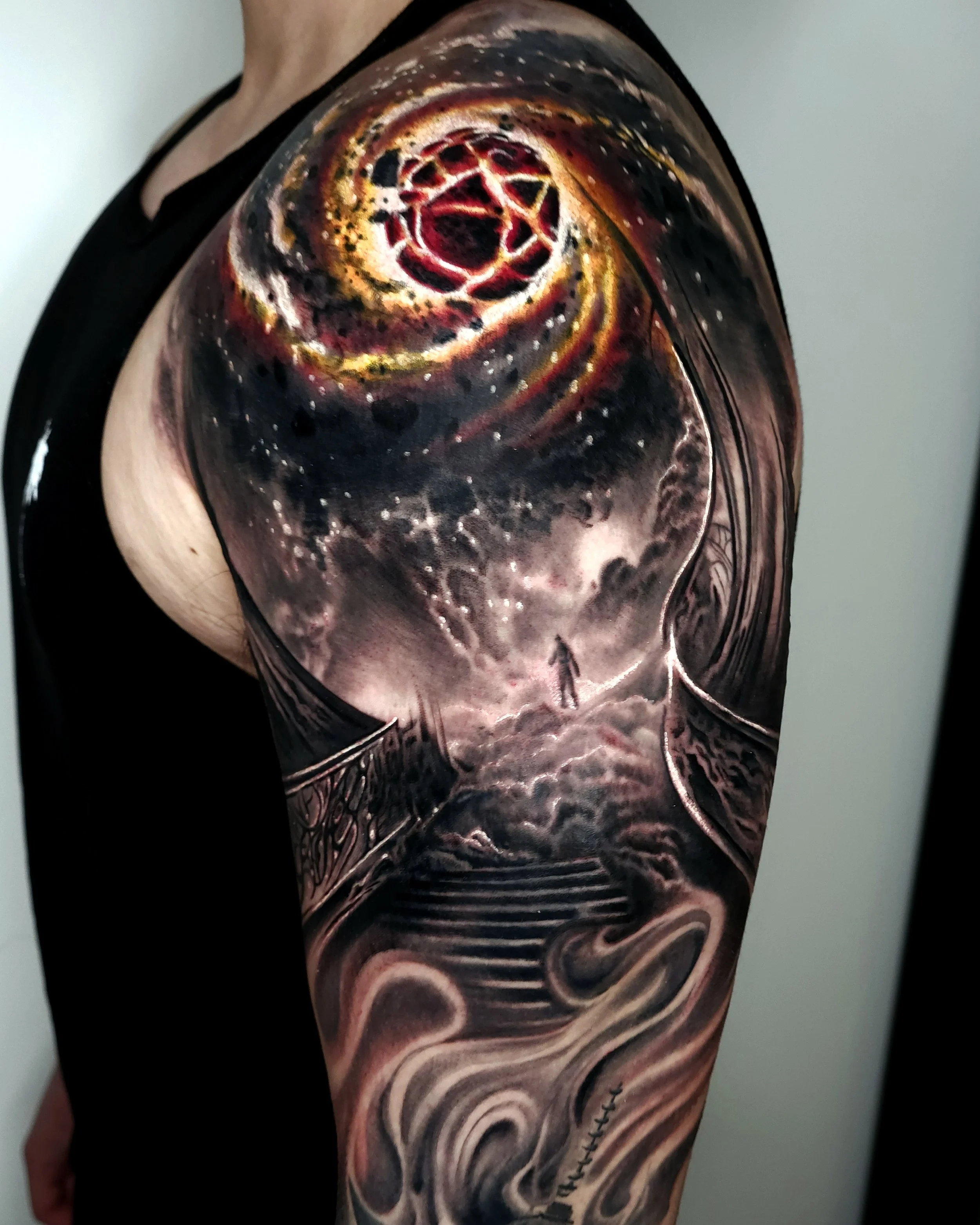 A detailed galaxy and space-themed tattoo on a person's upper arm, featuring a cosmic scene with swirling clouds, stars, and a tunnel-like formation leading to a figure standing on a cloud.