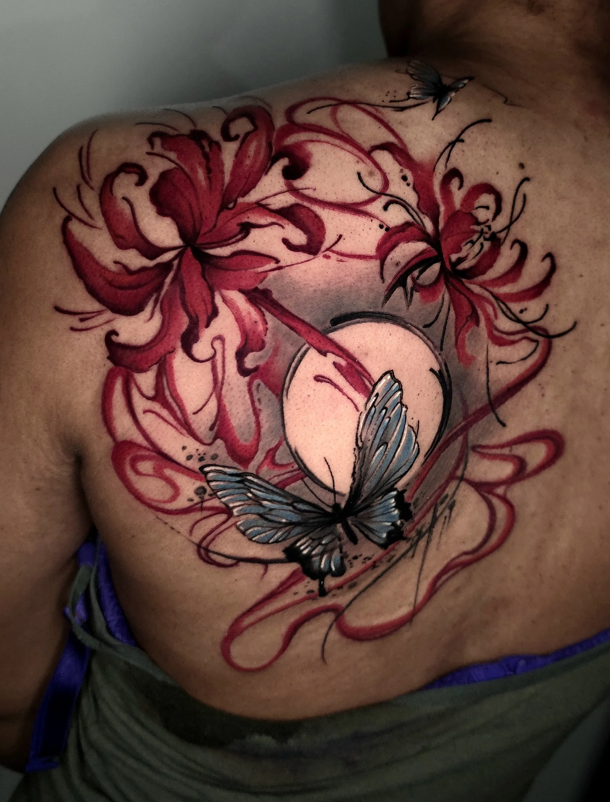 Tattoo of red lilies and two blue butterflies on a person's chest with a circular moon in the background.