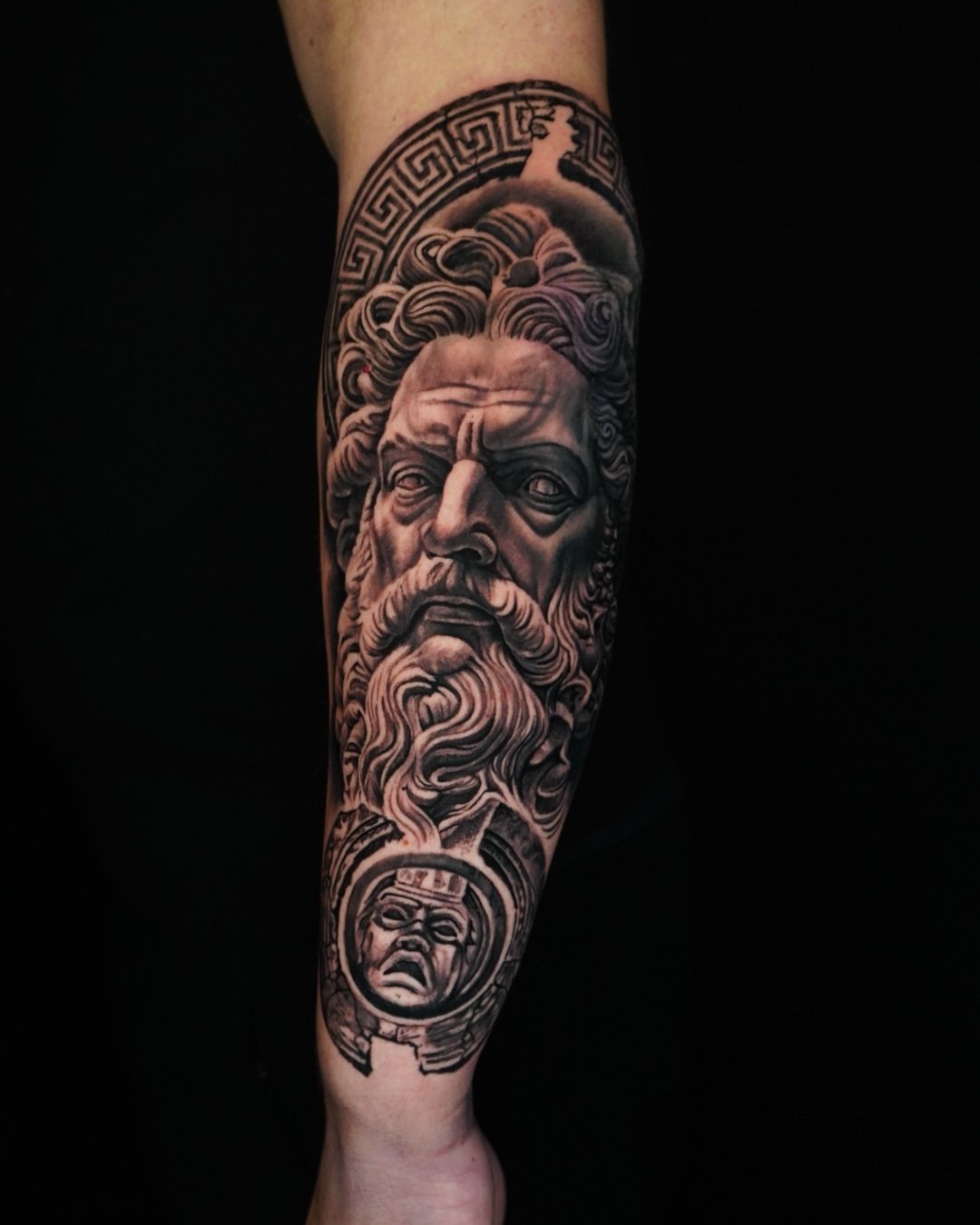 Black and gray tattoo of an ancient Greek or Roman bearded man with flowing hair on the forearm, with a circular design featuring a distressed face below the beard.