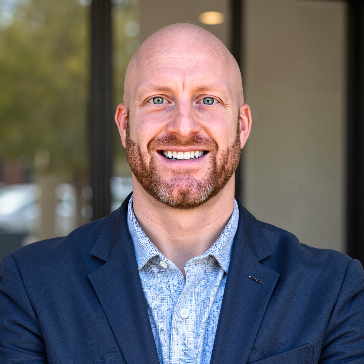 Headshot of a bald man with a beard smiling outdoors, wearing a navy blazer and a light patterned shirt.