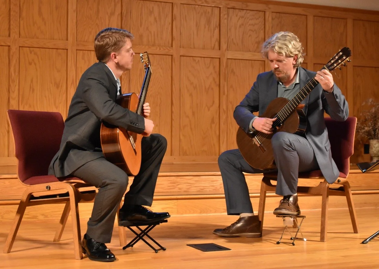 Two men dressed in suits sitting on chairs, playing acoustic guitars on a wooden stage with wood-paneled back wall.
