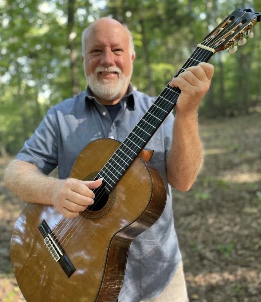 A man with a beard and short hair playing an acoustic guitar outdoors, surrounded by green trees.