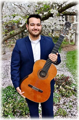 A young man in a blue suit holding an acoustic guitar, standing outdoors in front of blooming cherry blossom trees.