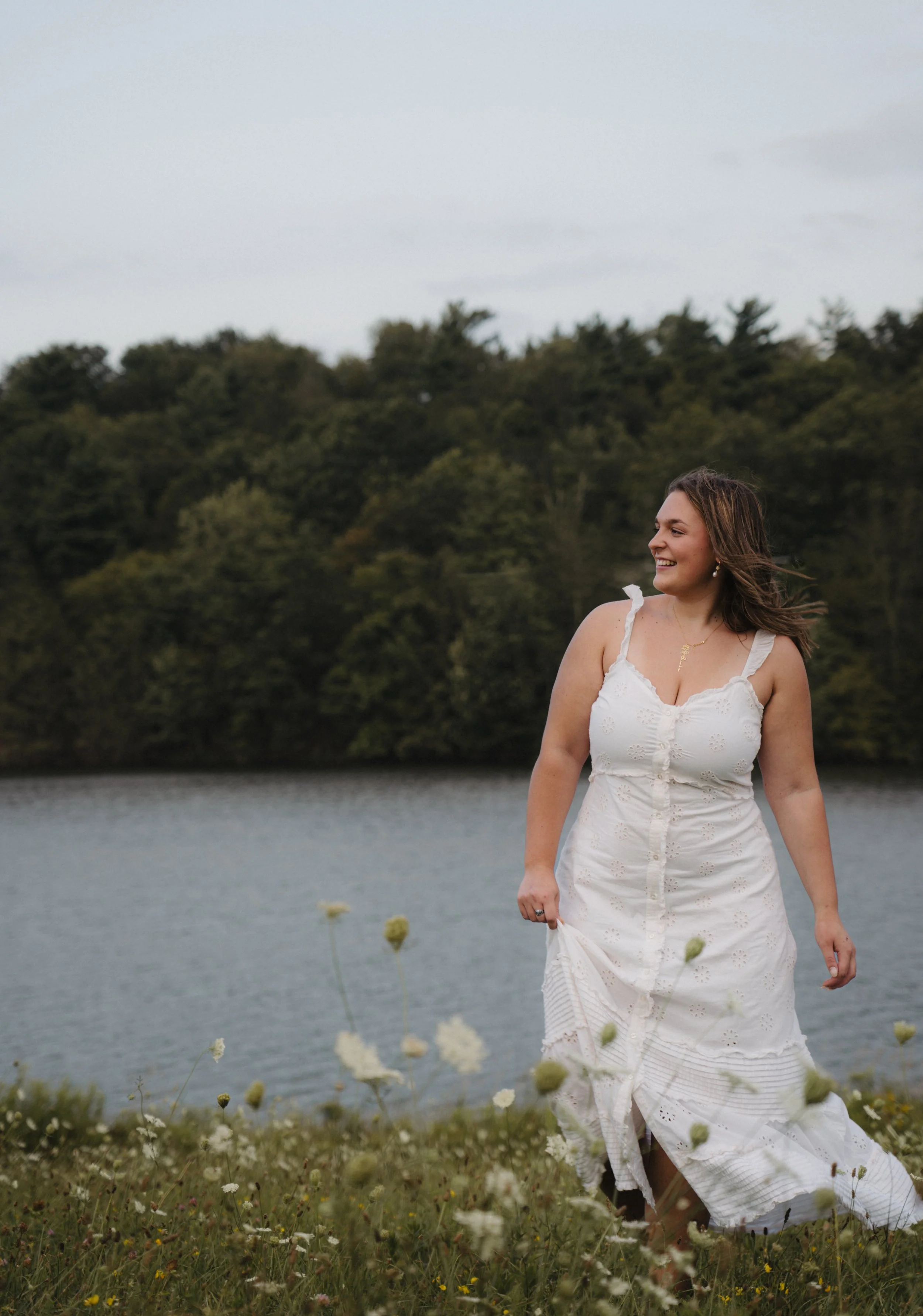 A woman in a white summer dress standing in a field of flowers near a lake with a background of trees and an overcast sky, smiling and looking to her right.