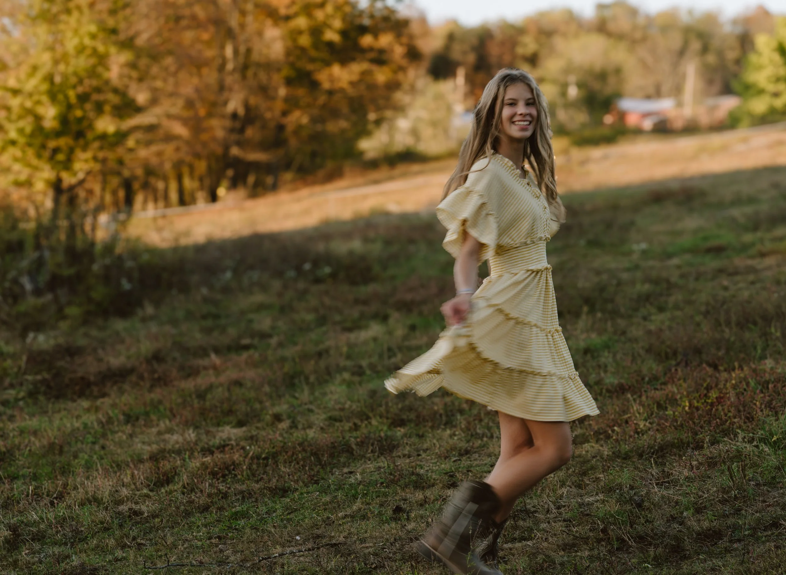 A young woman in a yellow dress and cowboy boots is smiling and spinning in a grassy field during autumn, with trees and a farm in the background.