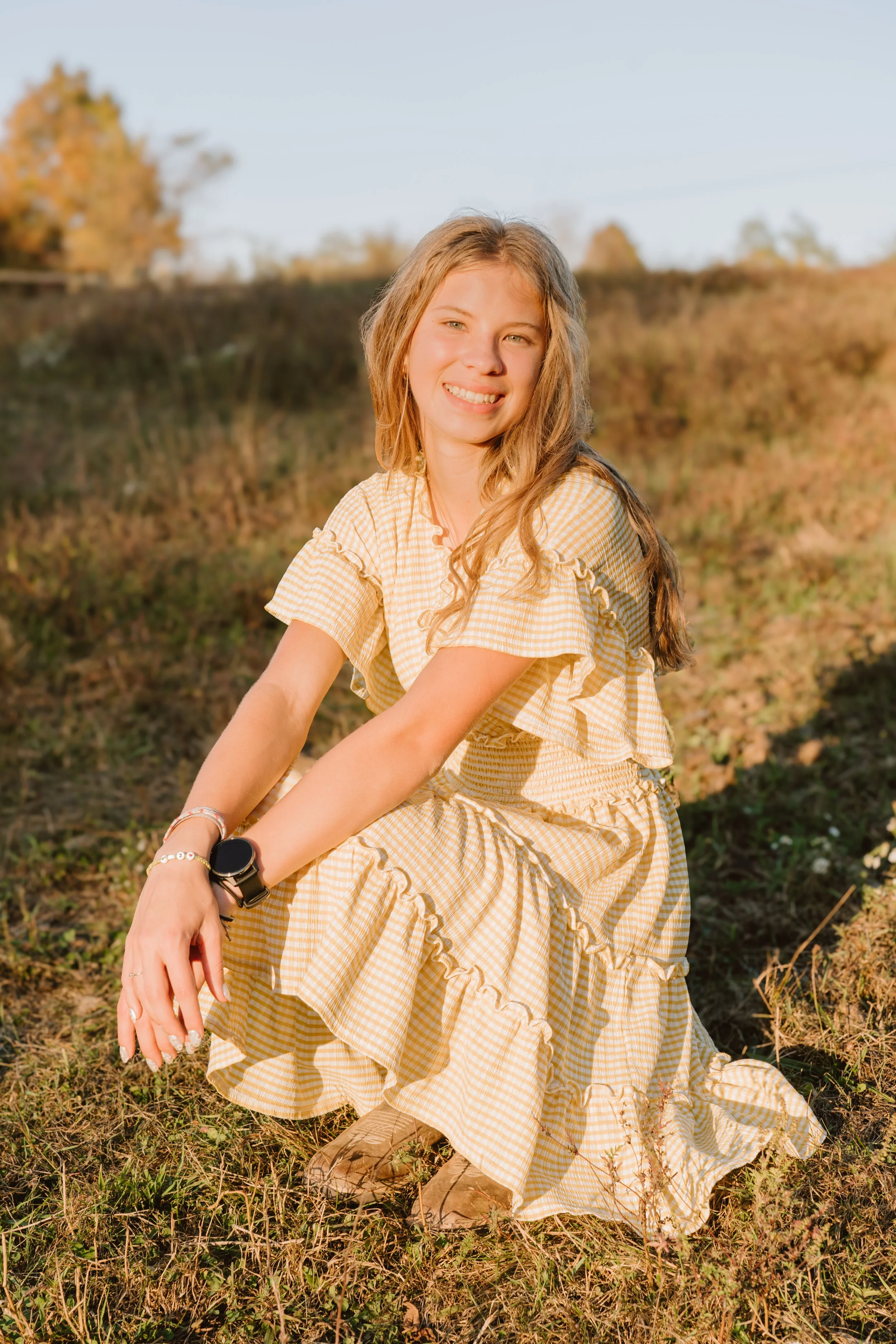 A young woman with long blonde hair sitting on the ground outdoors in a field during sunset, wearing a yellow checkered dress and smiling at the camera.