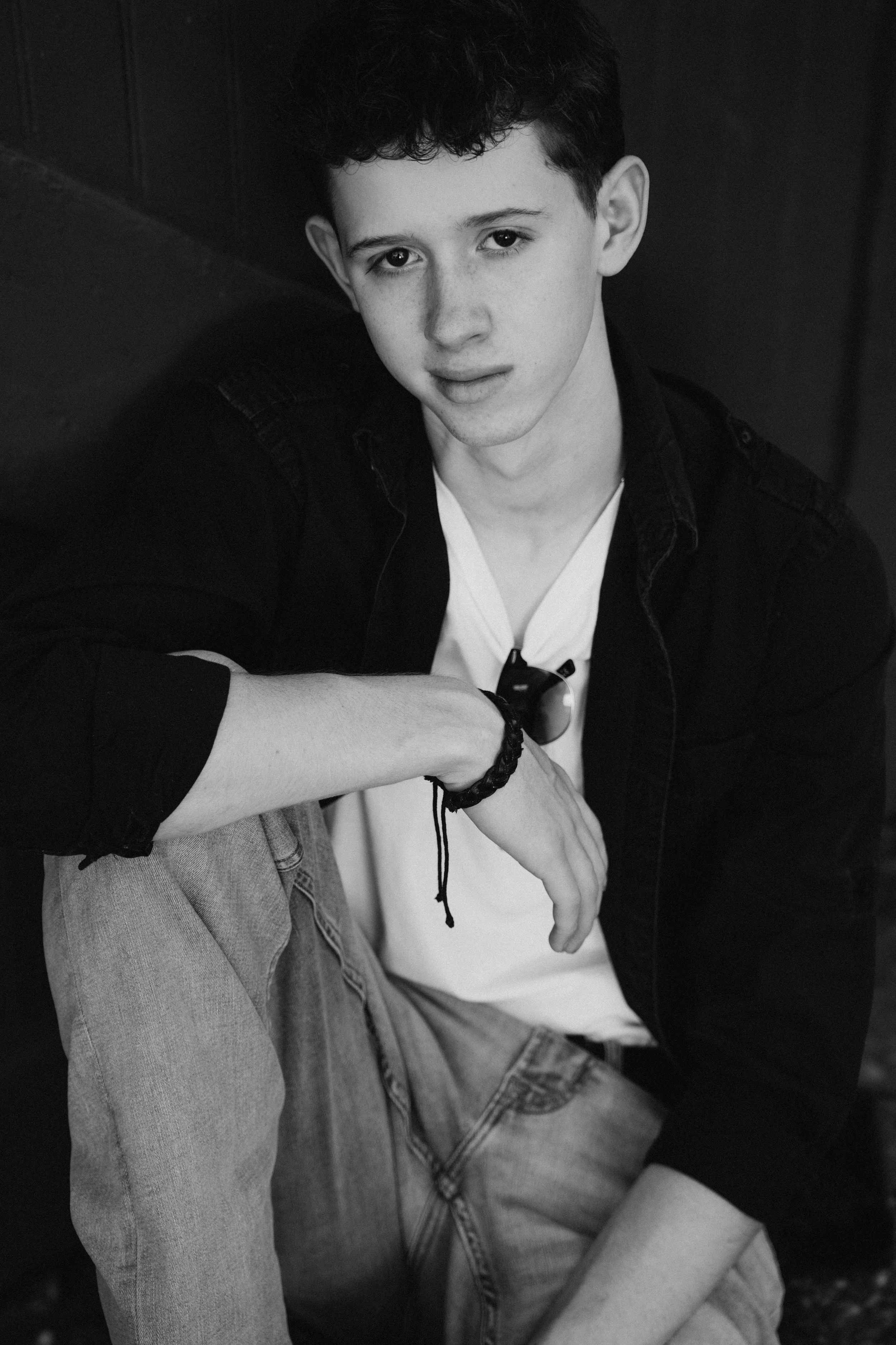 A young man with curly hair wearing a white T-shirt, black denim jacket, and light-washed jeans, sitting against a dark wall and looking at the camera.