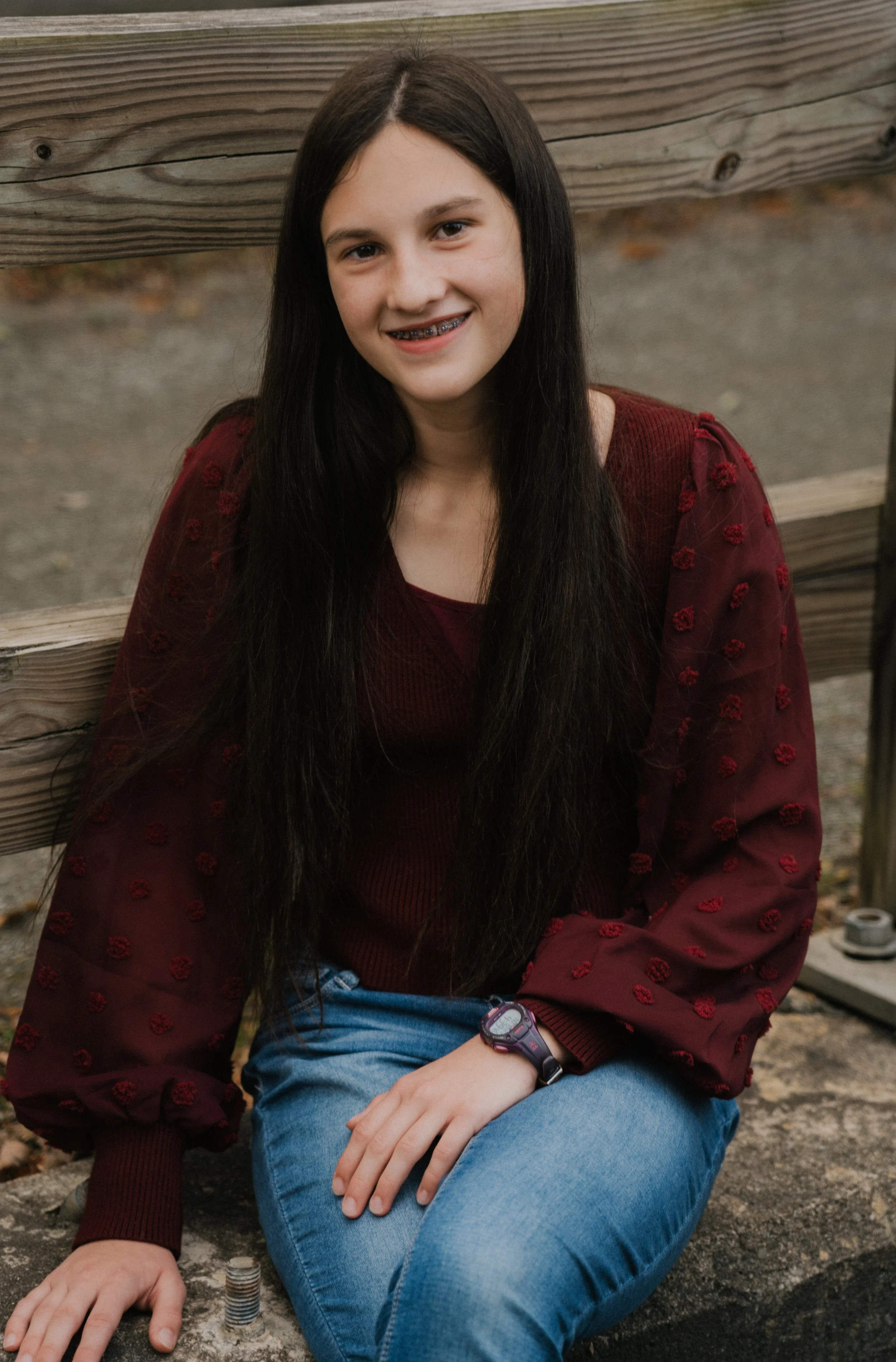 A young girl with long dark hair, wearing a maroon sweater with sheer embroidered sleeves, blue jeans, a watch, and braces, sitting outdoors near a wooden fence and smiling.
