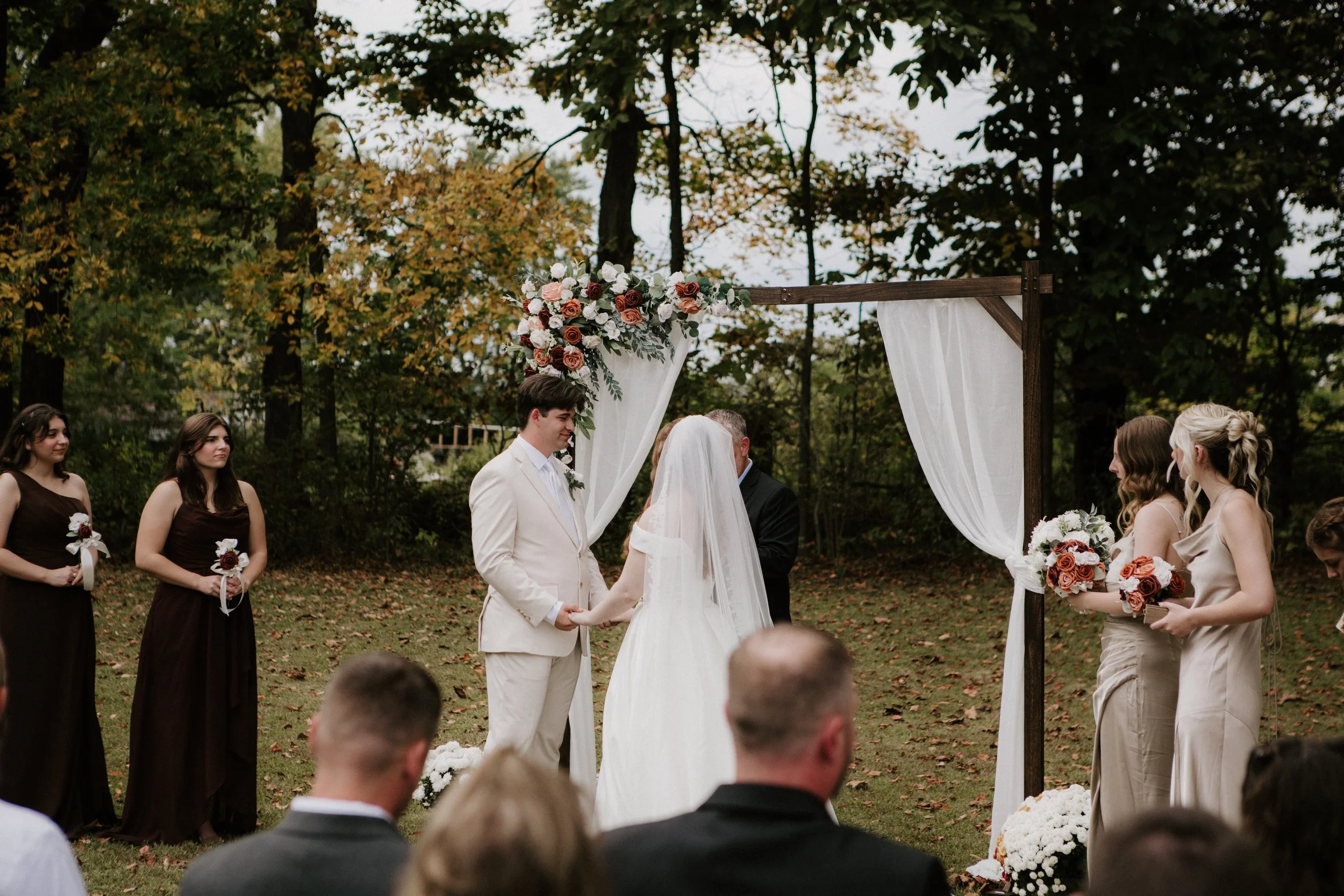 A wedding ceremony outdoors with a couple holding hands and exchanging vows, standing under a wooden arch decorated with white drapes and a floral arrangement. Bridesmaids and groomsmen hold bouquets and look on as guests watch.