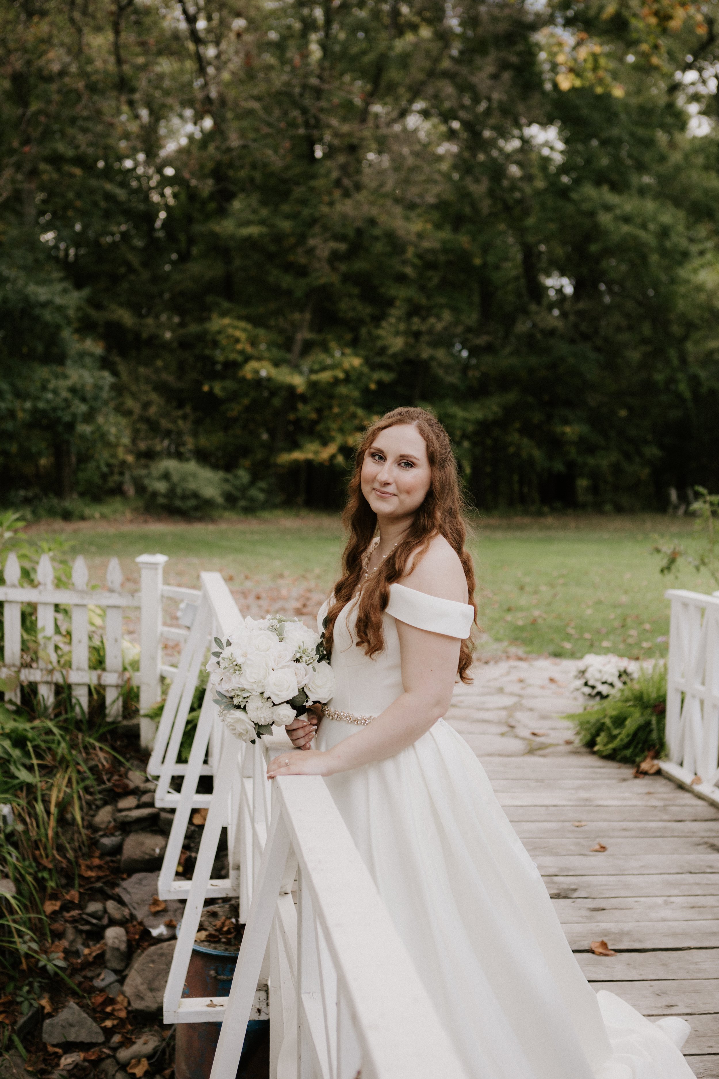 A bride with long, curly red hair in a white off-shoulder wedding gown holding a bouquet of white roses on a white wooden bridge outdoors, with trees and greenery in the background.
