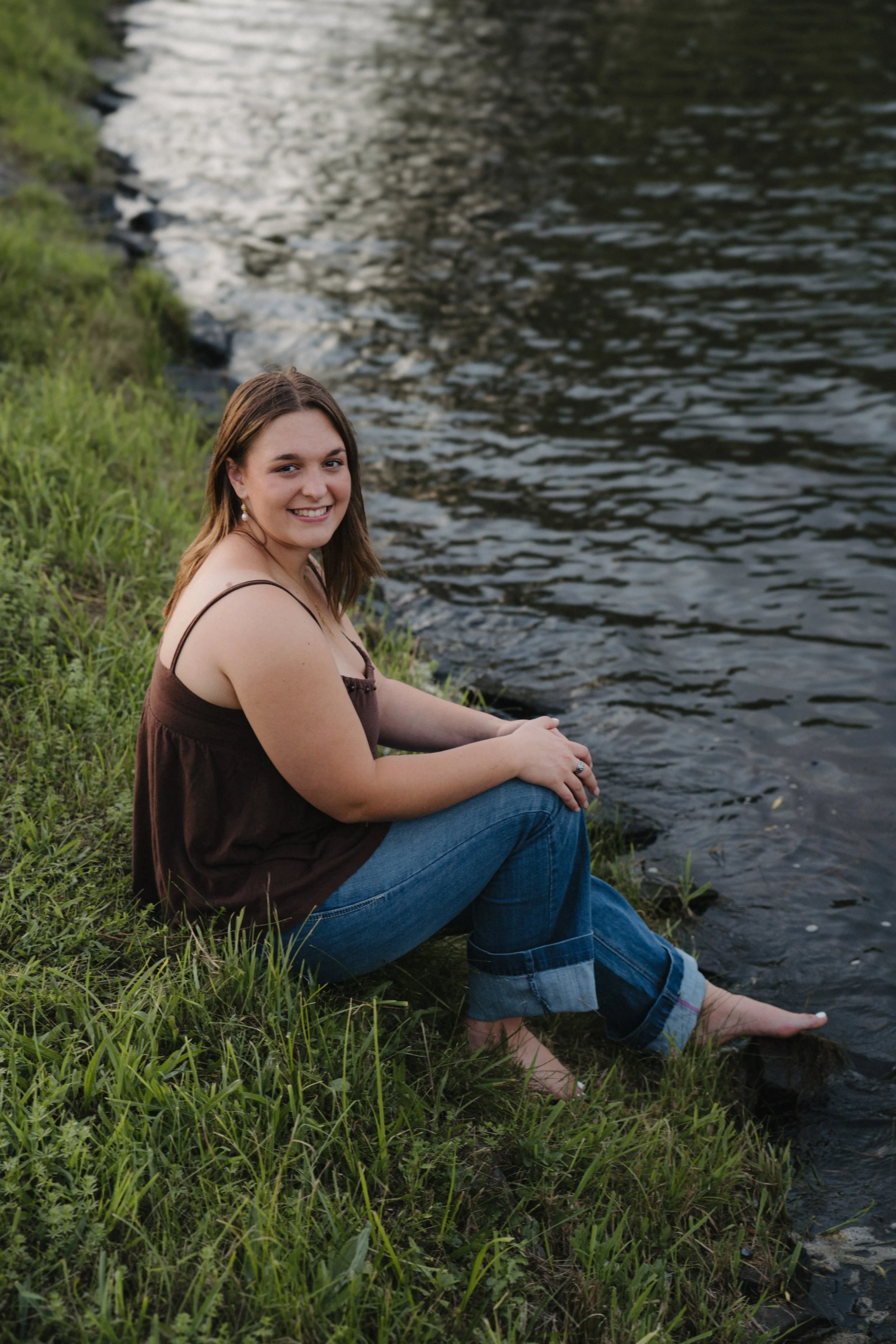 Young woman sitting by a lake with green grass, smiling, wearing a brown tank top and blue jeans with rolled-up cuffs.