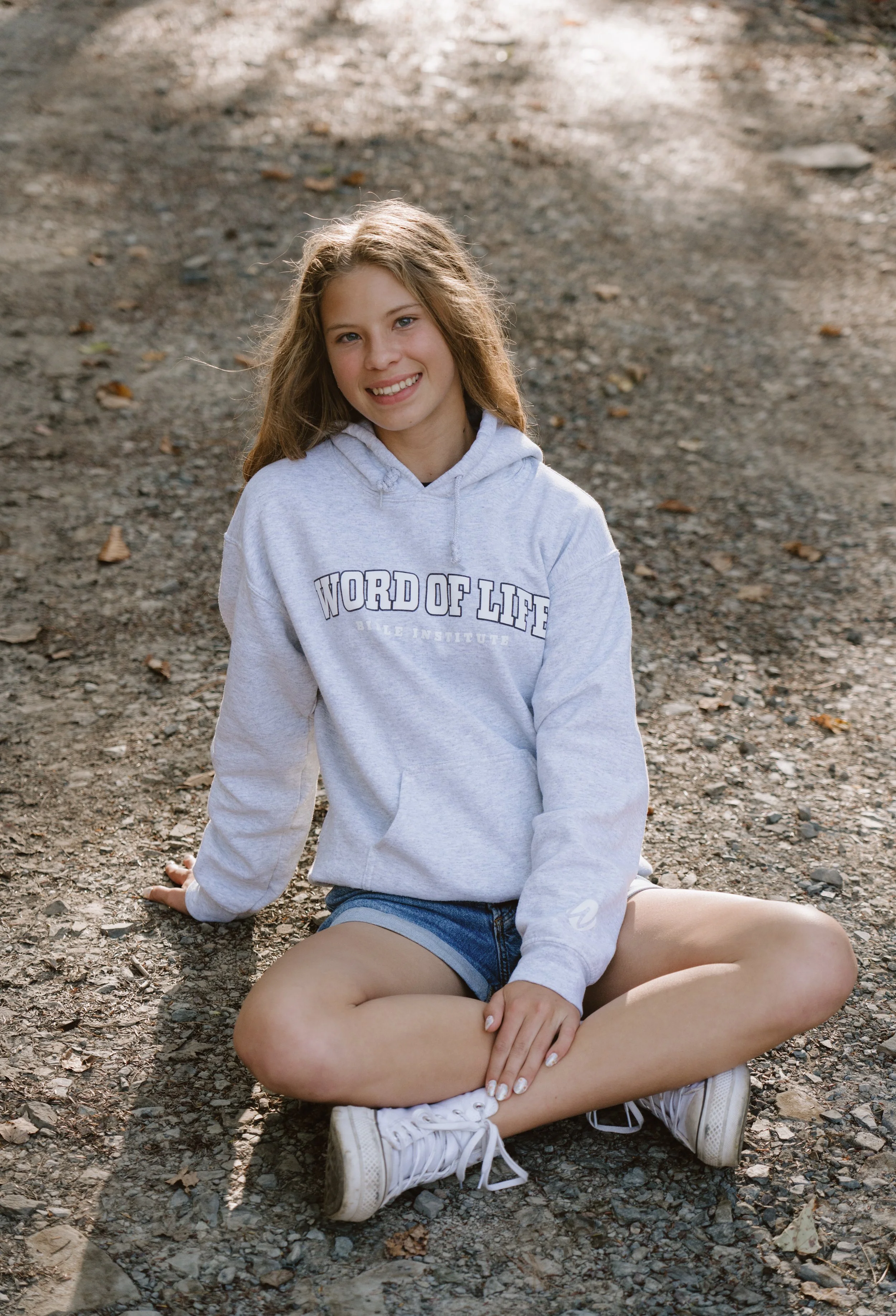 A young girl with long light brown hair, smiling, sitting cross-legged on a gravel path outdoors. She is wearing a gray hoodie with the words 'Word of Life' and denim shorts.