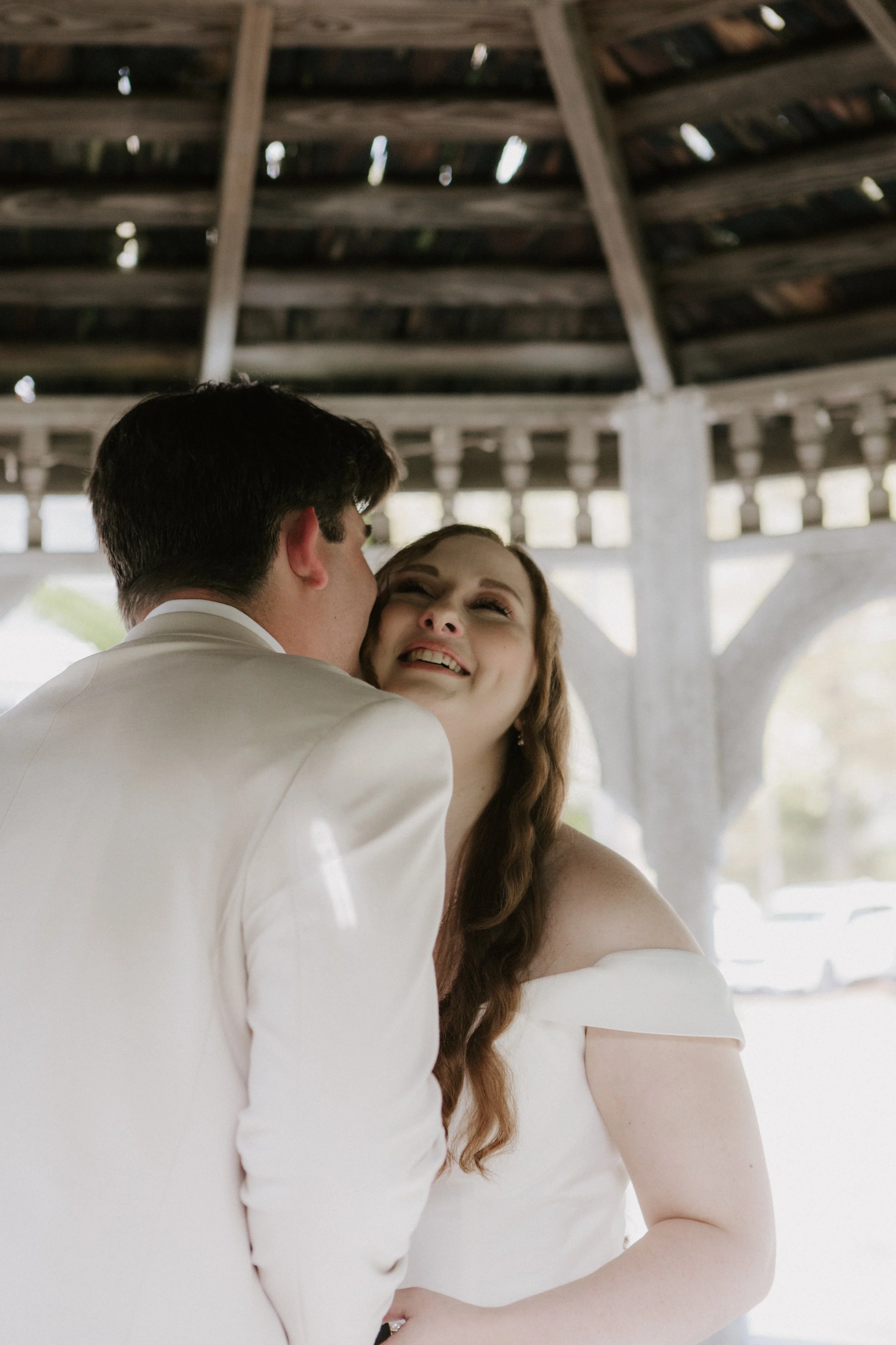 A bride and groom share a kiss under a rustic wooden pavilion during their wedding.