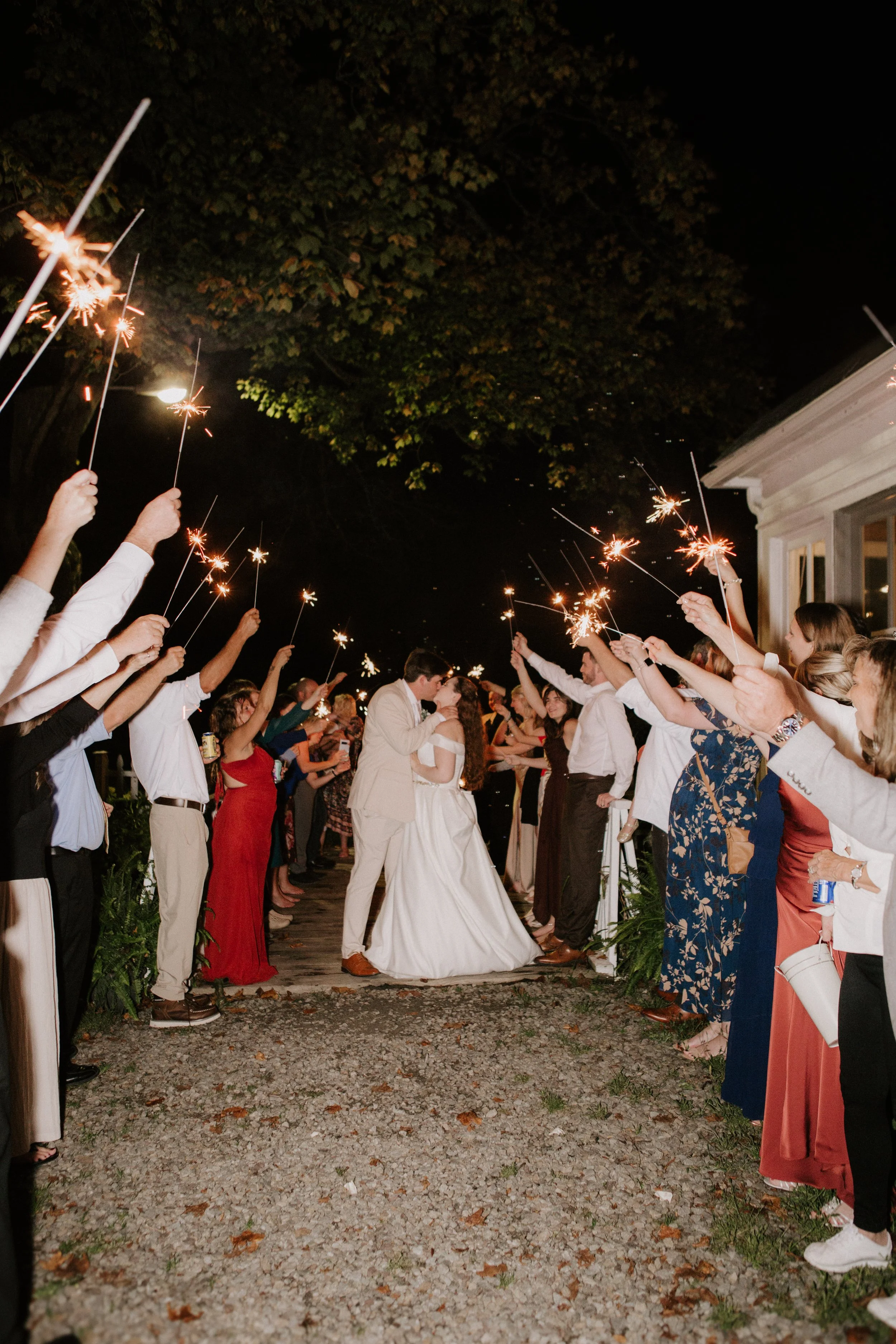 A bride and groom kiss during their wedding reception at night, surrounded by guests holding sparklers.