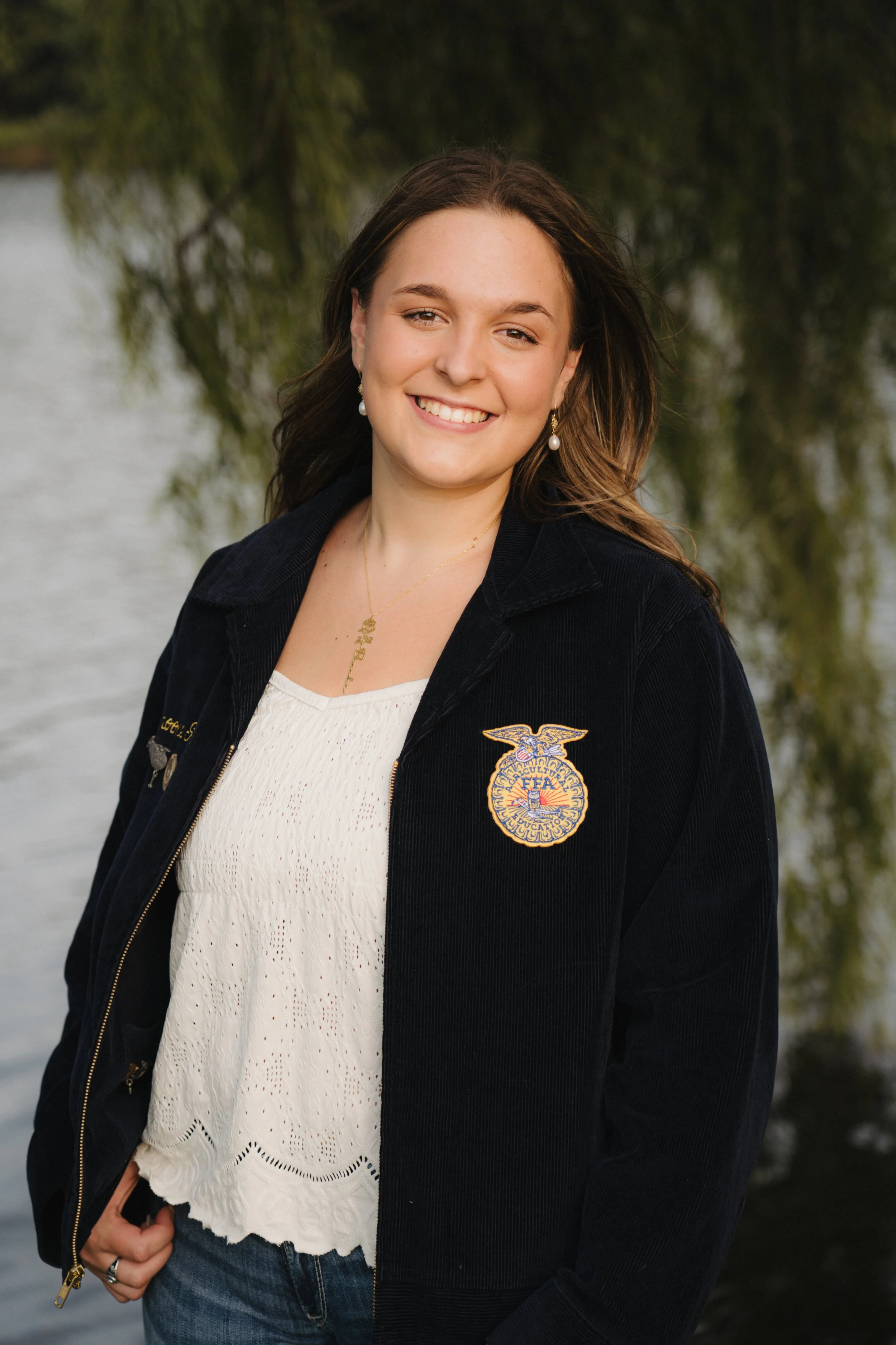 A young woman smiling outdoors next to water, wearing a black FFA jacket, white blouse, jeans, pearl earrings, and a gold necklace.