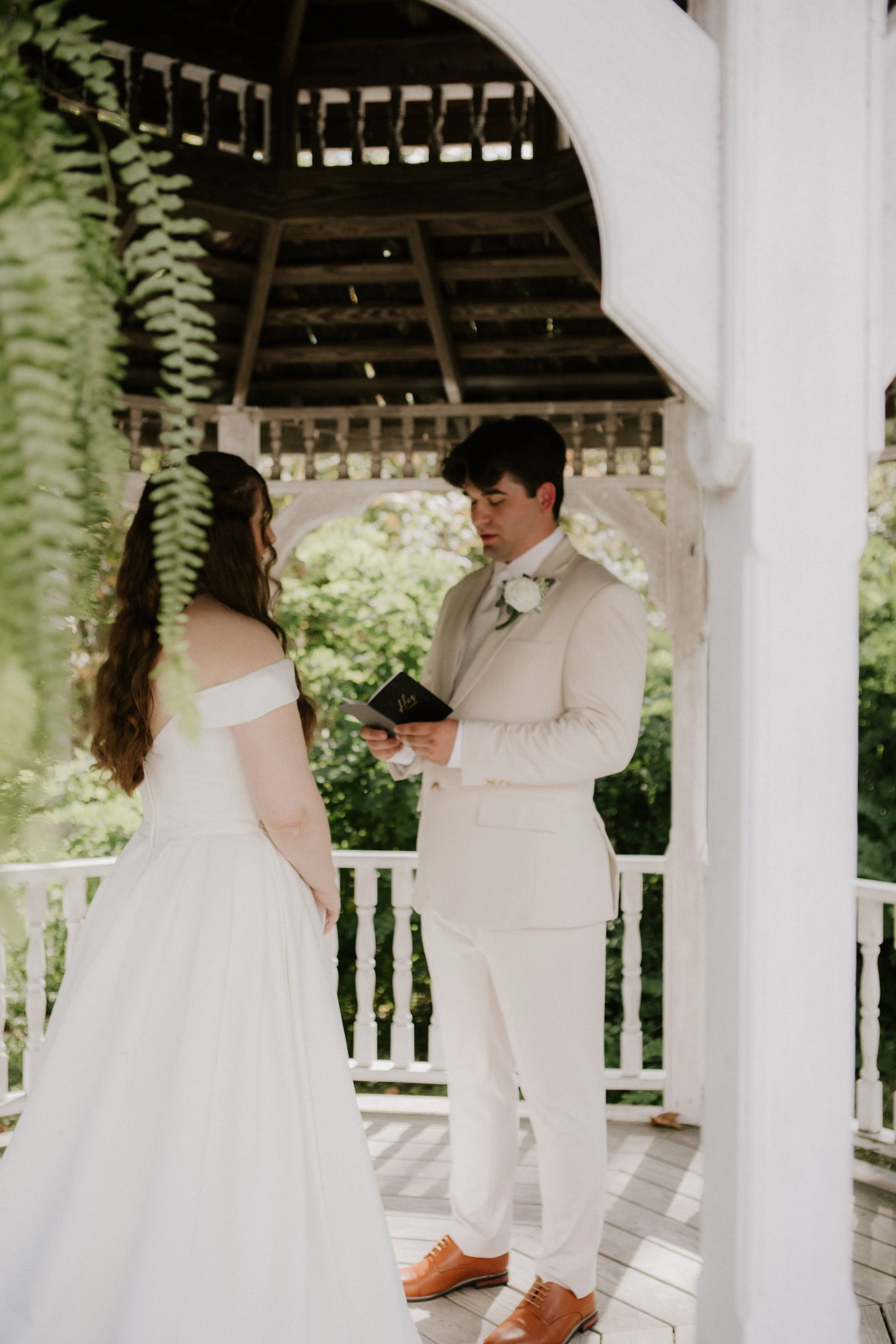 A couple getting married in an outdoor wedding ceremony under a white gazebo. The bride wears a white off-shoulder gown, and the groom is dressed in a white suit with brown shoes. The groom is reading vows from a small book.