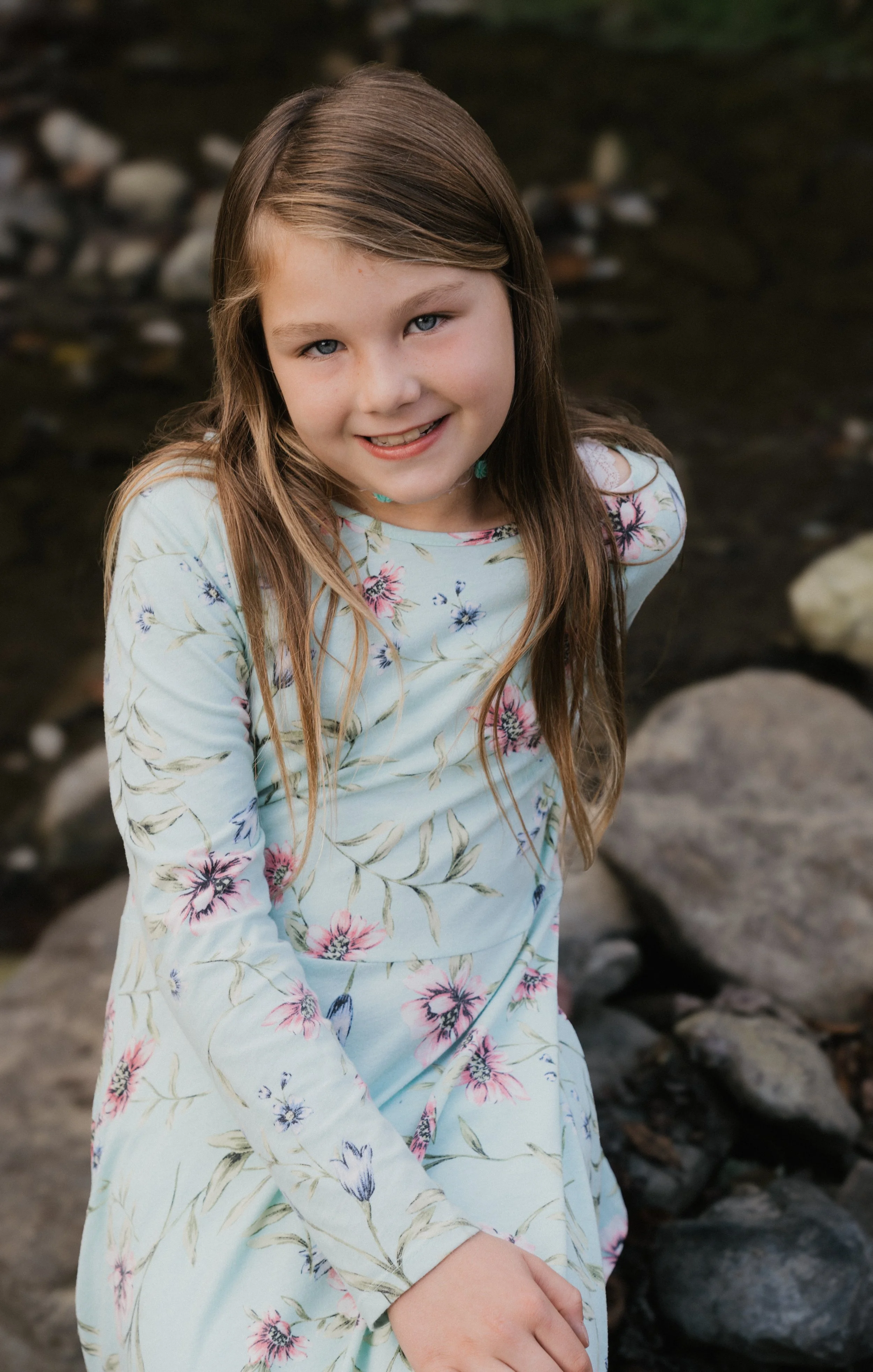 A young girl with long brown hair and blue eyes, wearing a light blue floral dress, sitting on rocks near a body of water, smiling at the camera.